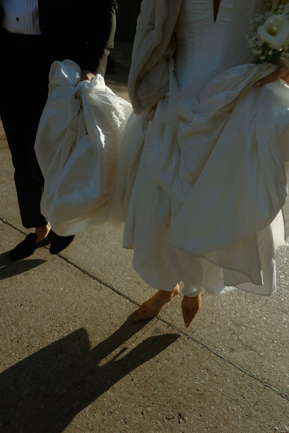 A white wedding dress and dress shoes cast shadows on concrete as someone in black stands nearby.