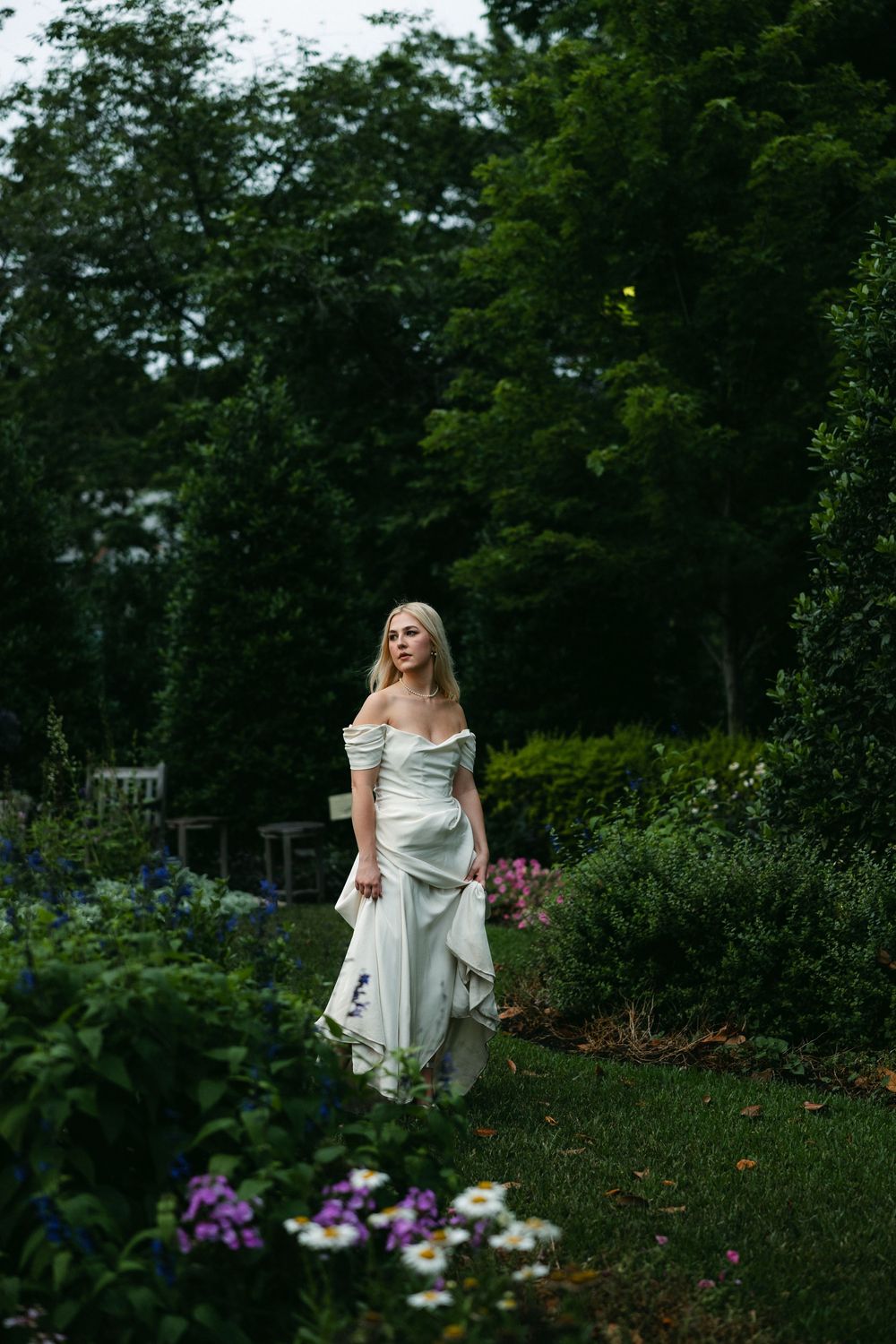 Bride in white dress walking through lush garden setting with purple flowers and green foliage.
