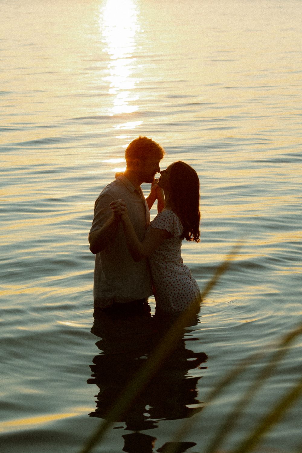 Silhouetted couple embraces while sitting in calm water at sunset creating a romantic scene against golden reflections.