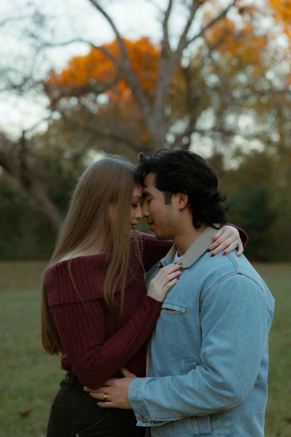 A romantic couple embraces and shares a kiss outdoors during autumn with orange leaves in the background.
