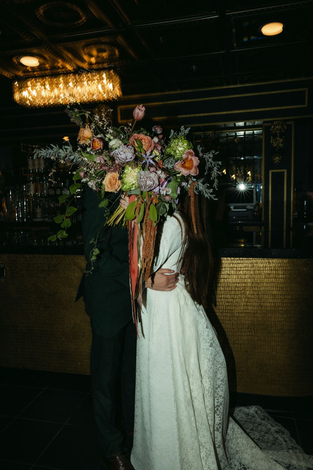 Wedding couple illuminated by overhead lights in dark reception venue with floral decorations.