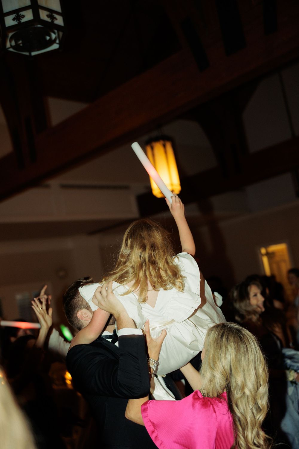People dancing and celebrating at a dimly lit wedding reception with warm glowing lights in the background.