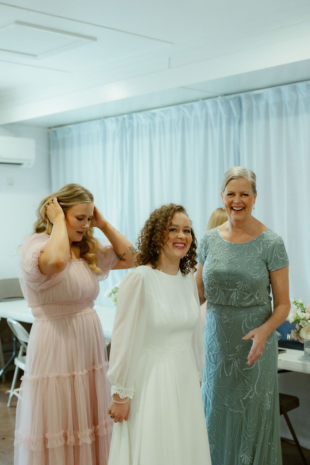 A group of women in formal dresses laugh together in a bright white room during a special occasion.
