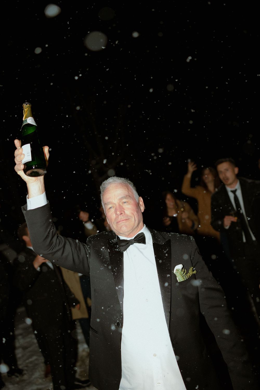 A person in a tuxedo holds up a champagne bottle while celebrating at a nighttime event with falling snow or confetti.
