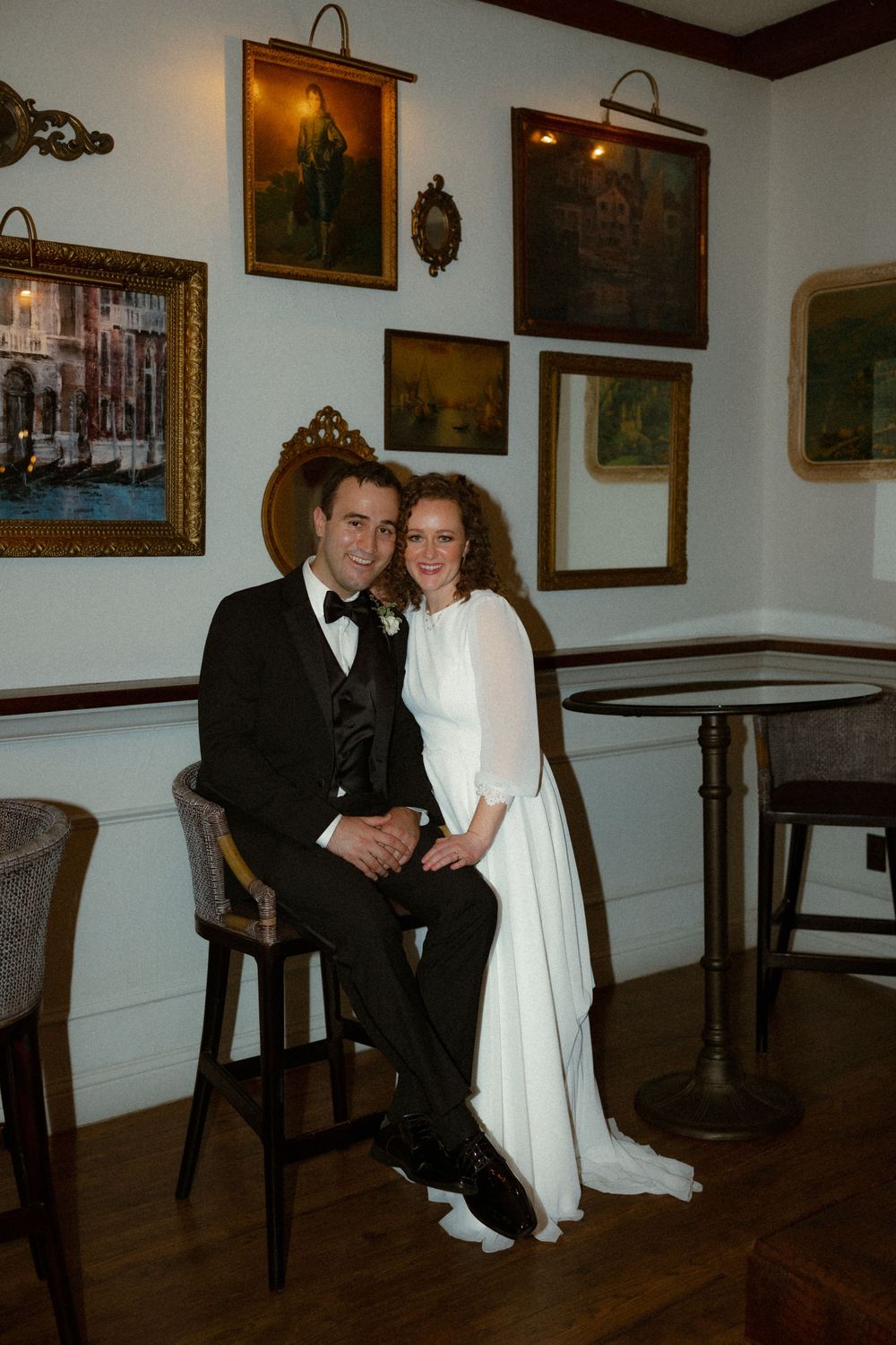 A couple in formal wedding attire poses together in a room decorated with vintage paintings and dark wood furniture.