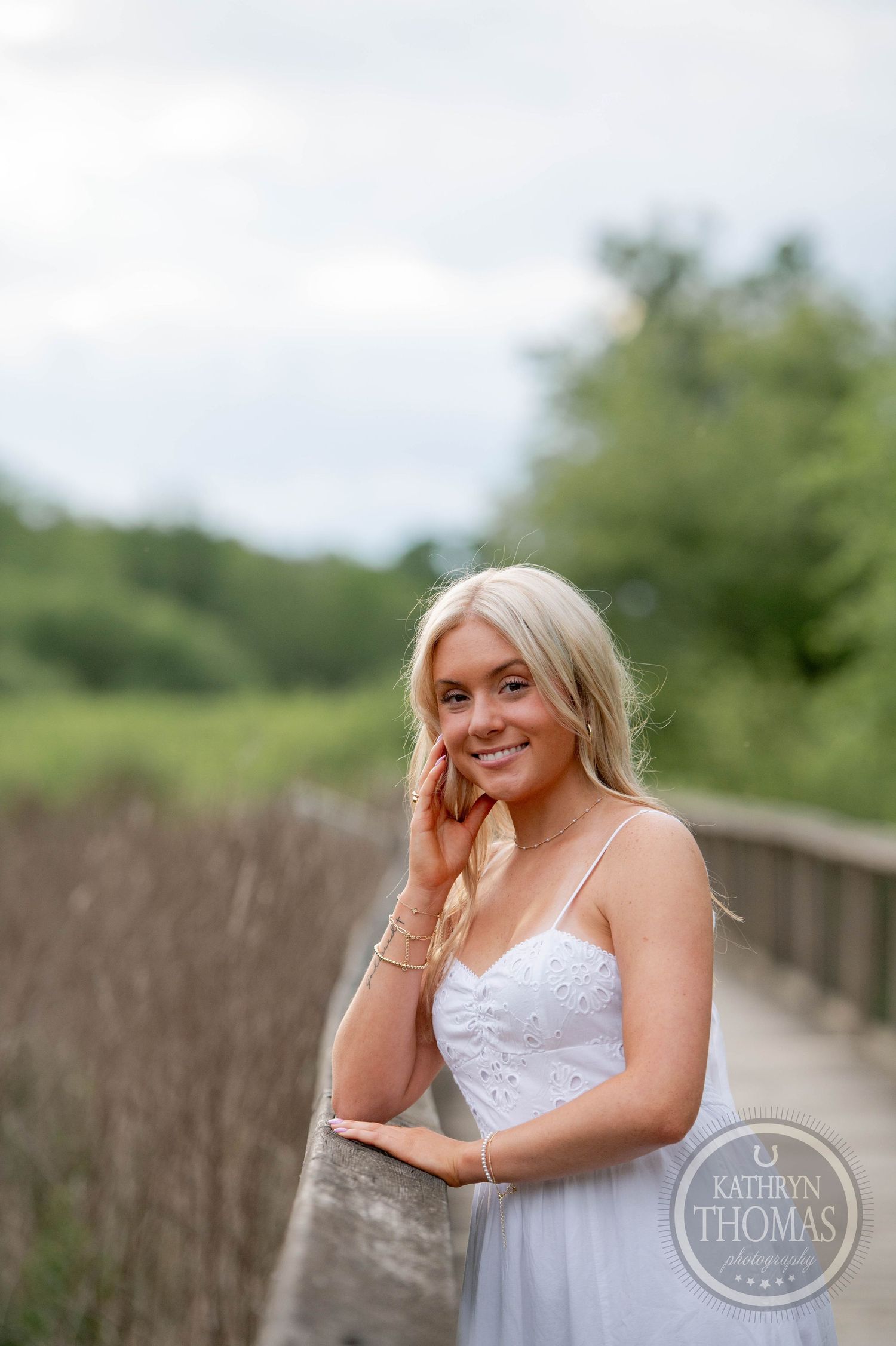 A person in a white spaghetti strap dress poses outdoors on a wooden bridge surrounded by greenery.