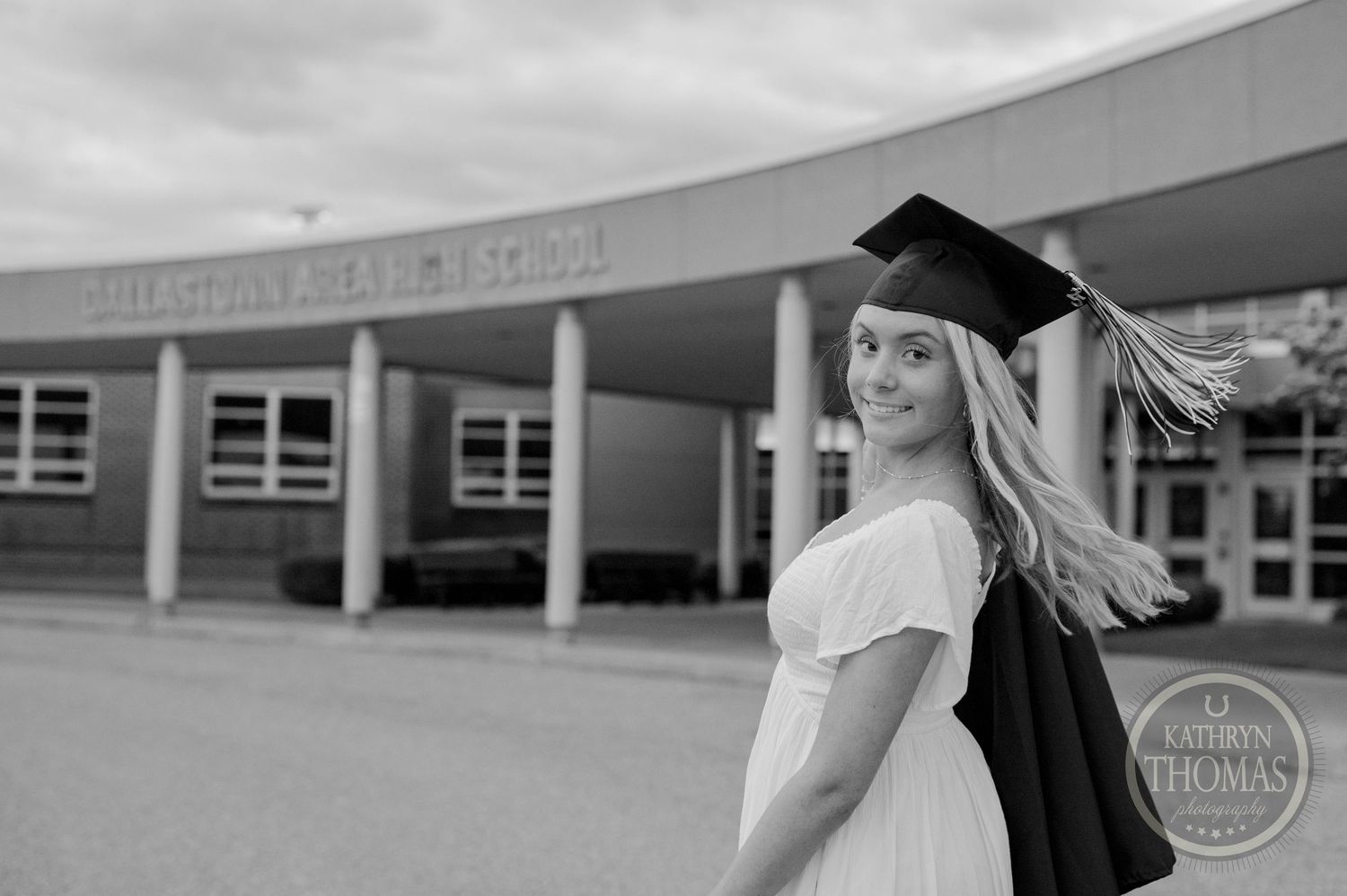 A graduate in cap and gown poses outside of a school building entrance in black and white photography.