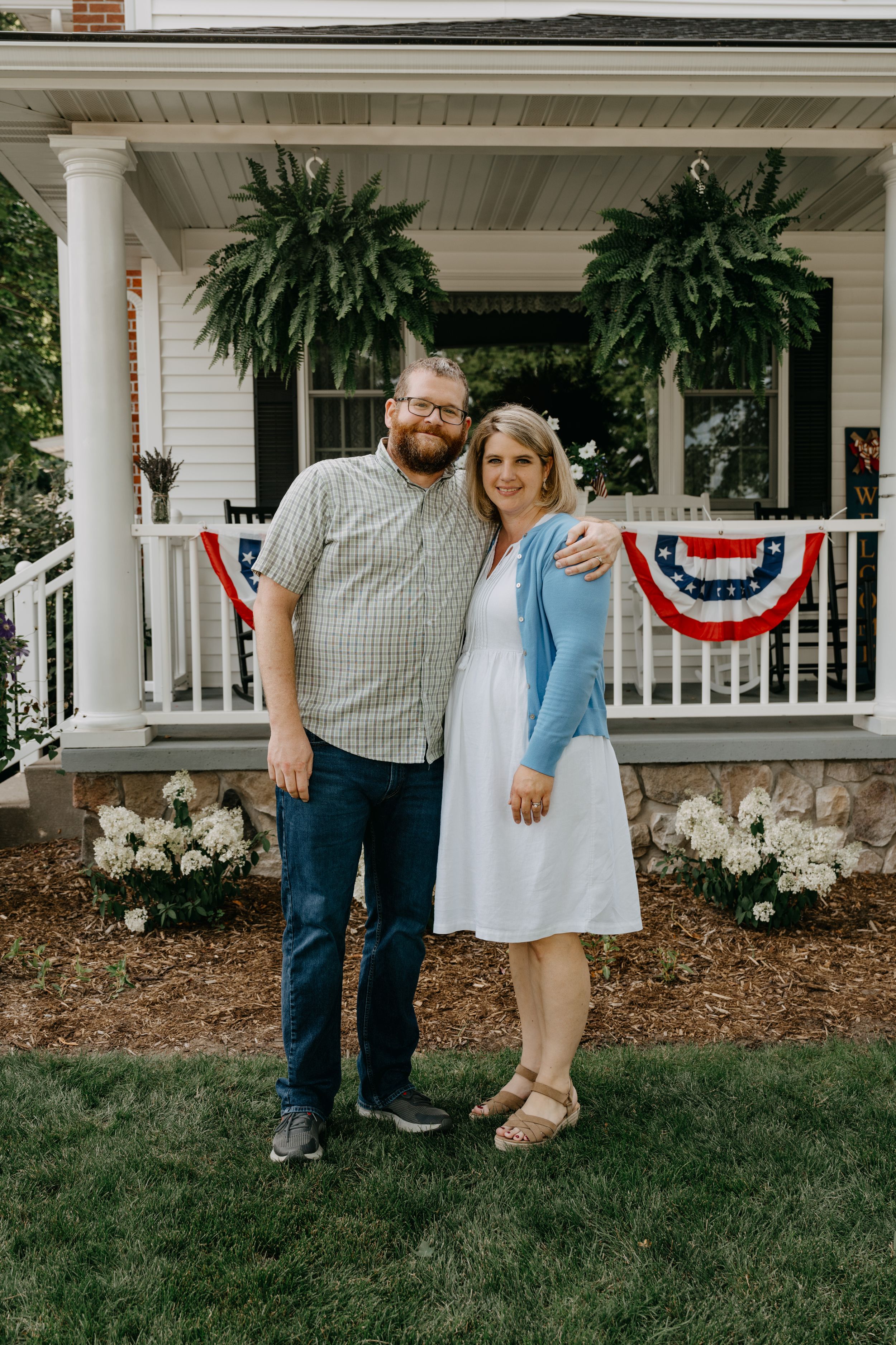 A couple stands together on the lawn in front of a classic white porch decorated with patriotic bunting.