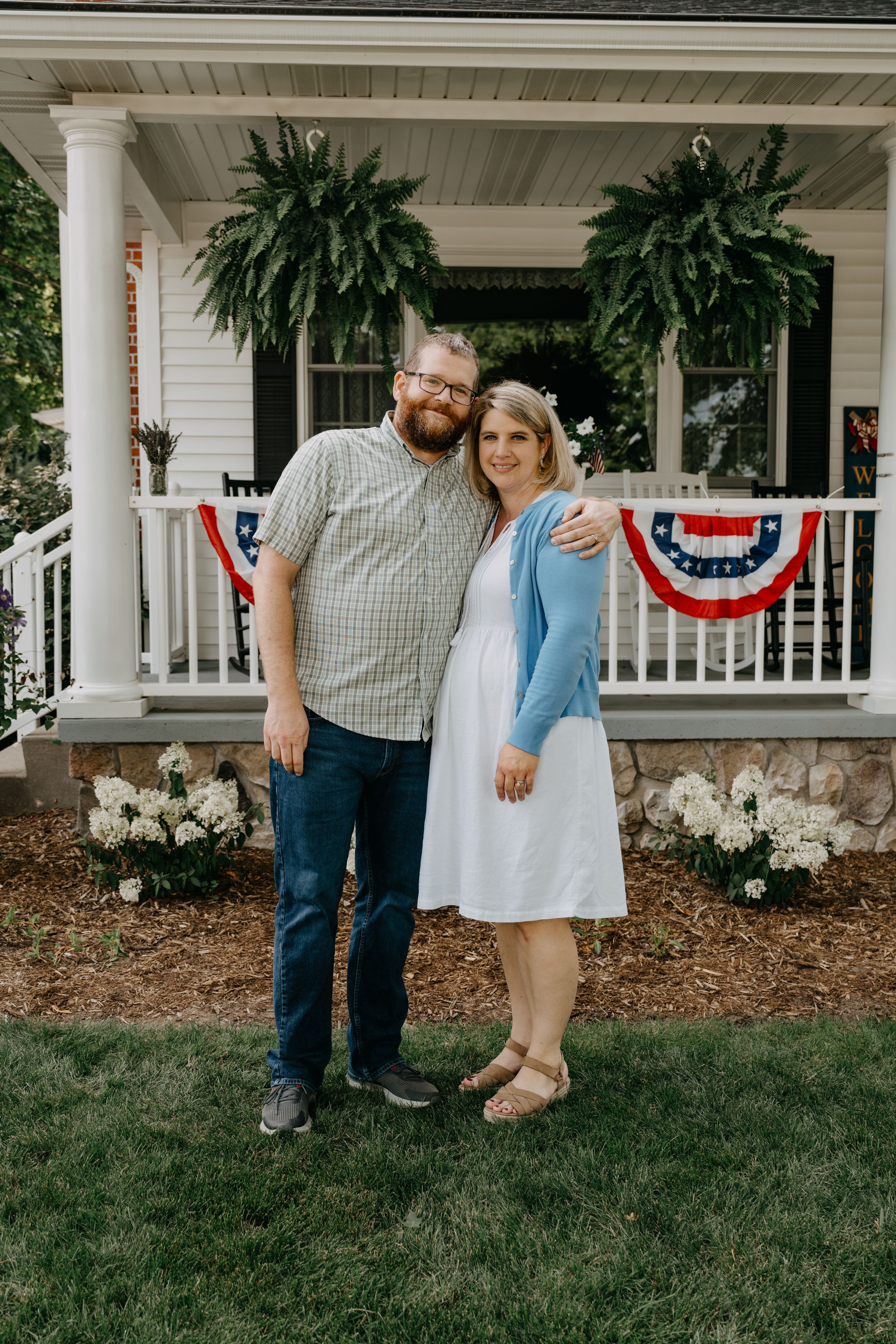 A couple stands on a front porch decorated with hanging ferns and patriotic bunting on a white house.