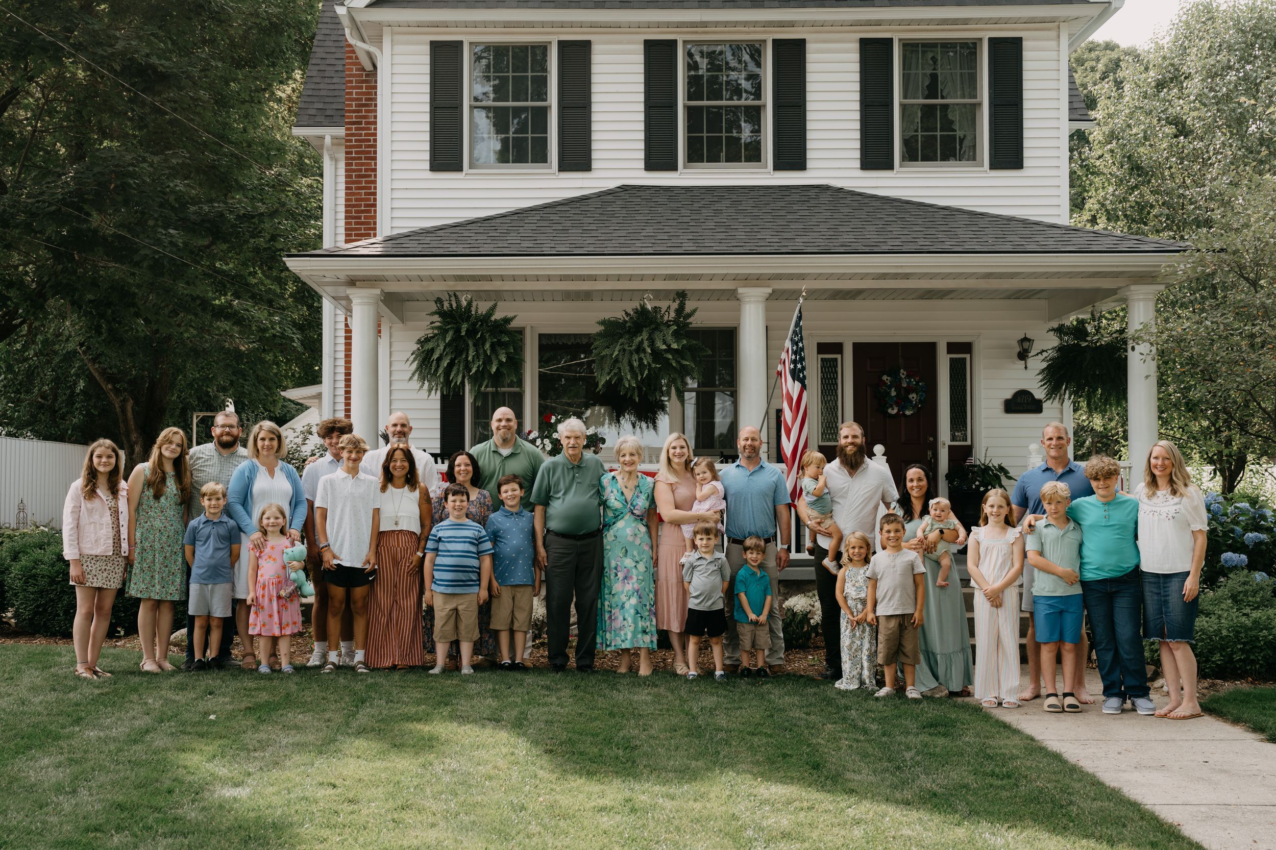 A large group of people gathered outside a white colonial-style house with a front porch for a summer celebration.