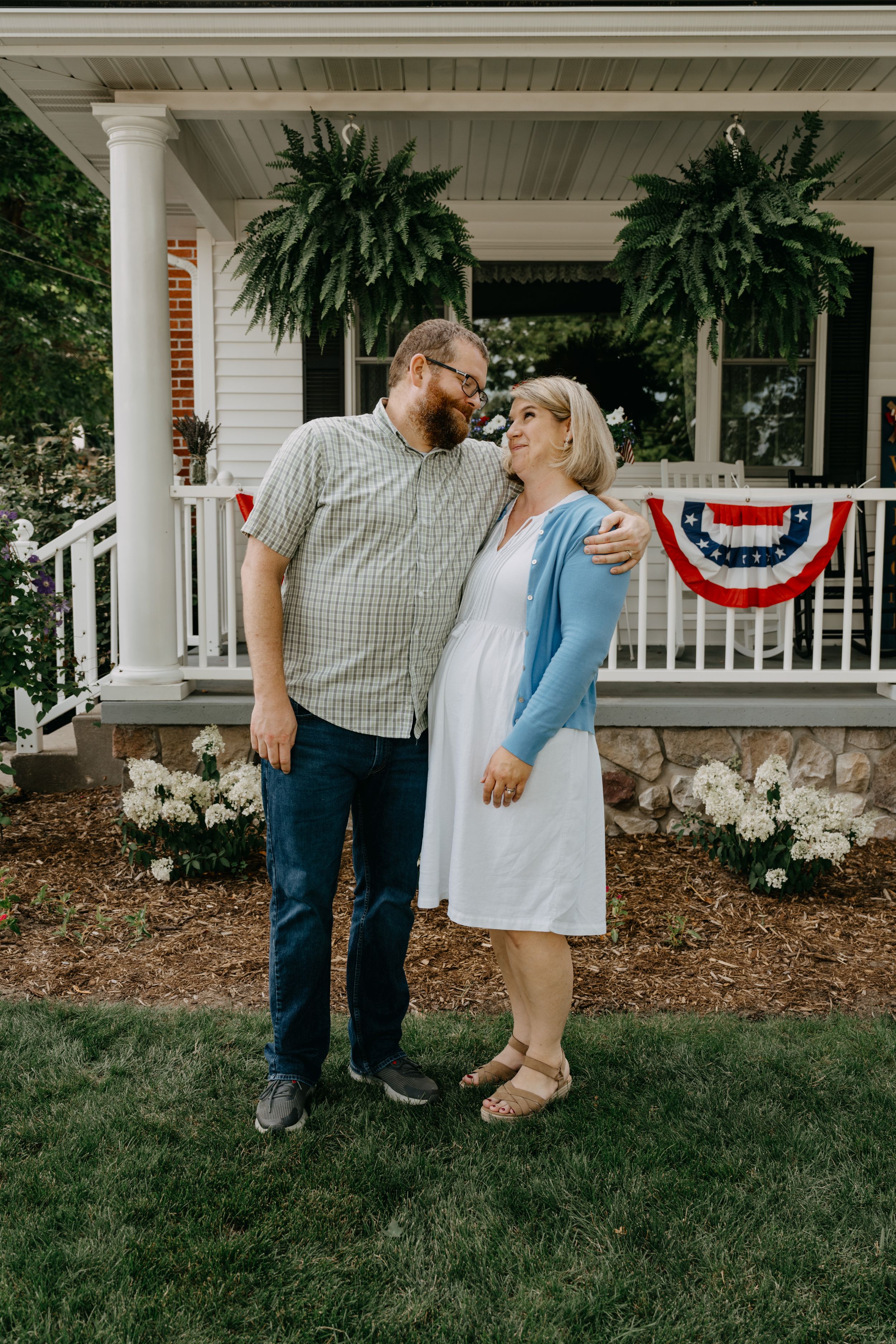 A sweet moment between a couple on their porch with patriotic decor and hanging ferns in the background.