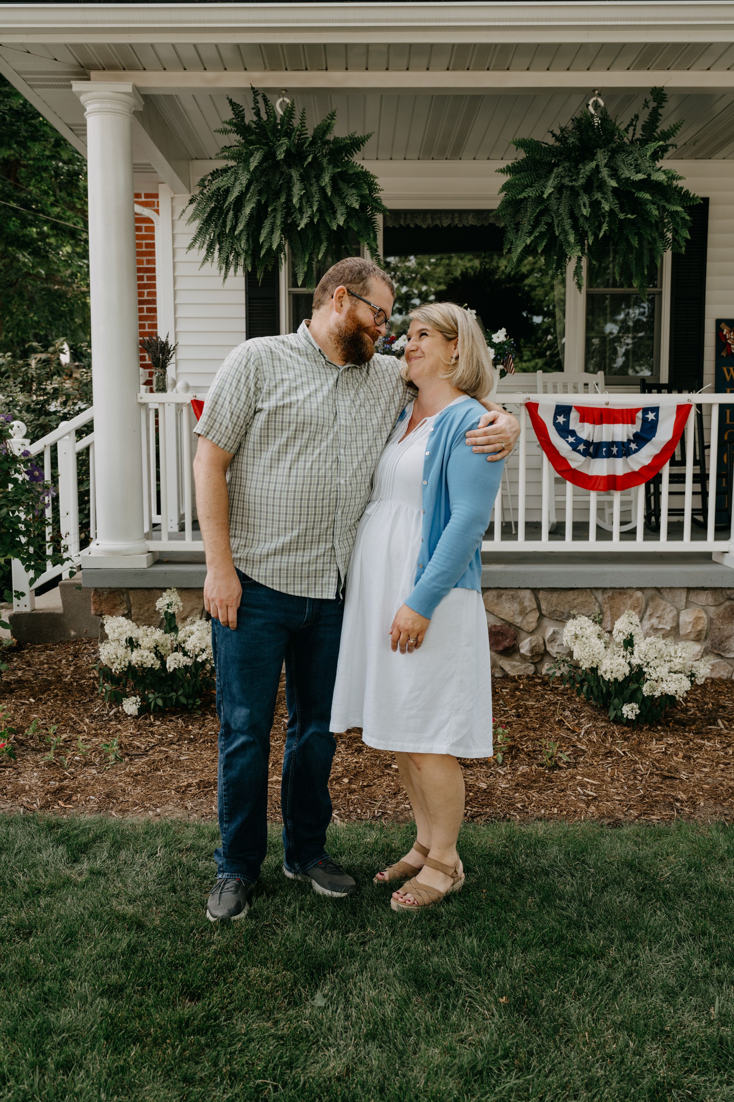 A romantic couple shares a sweet moment on a porch with patriotic decorations and hanging plants.