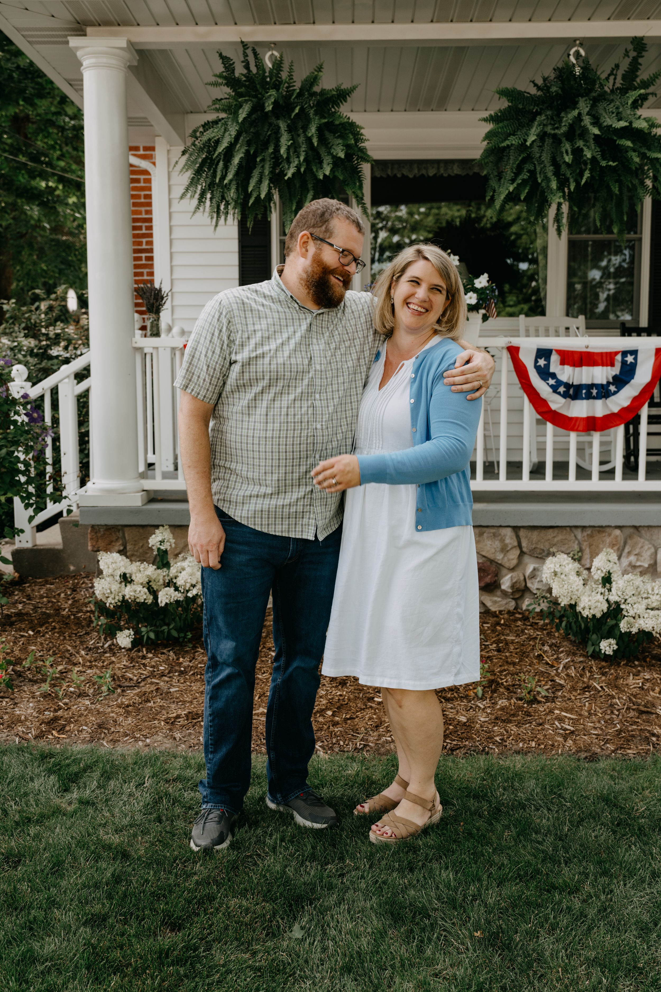 A couple embraces on their front lawn in front of a white porch adorned with hanging ferns and American bunting decor.
