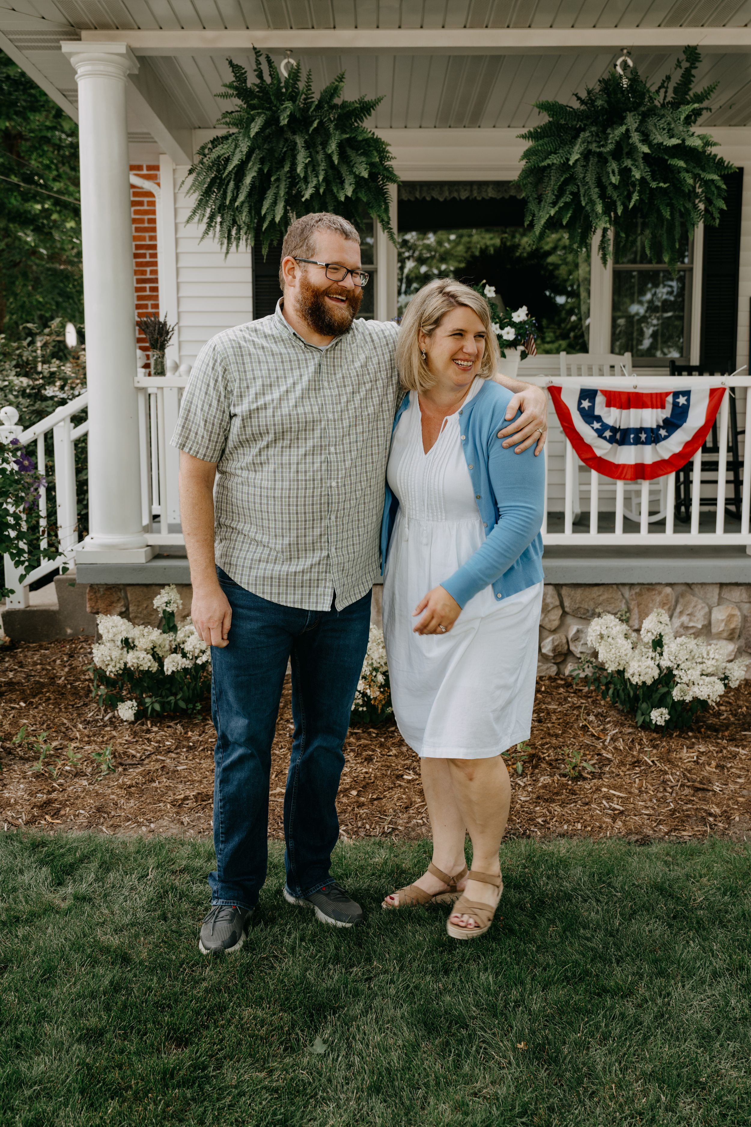 A couple stands together on a porch with hanging ferns and patriotic bunting, surrounded by a well-manicured garden.