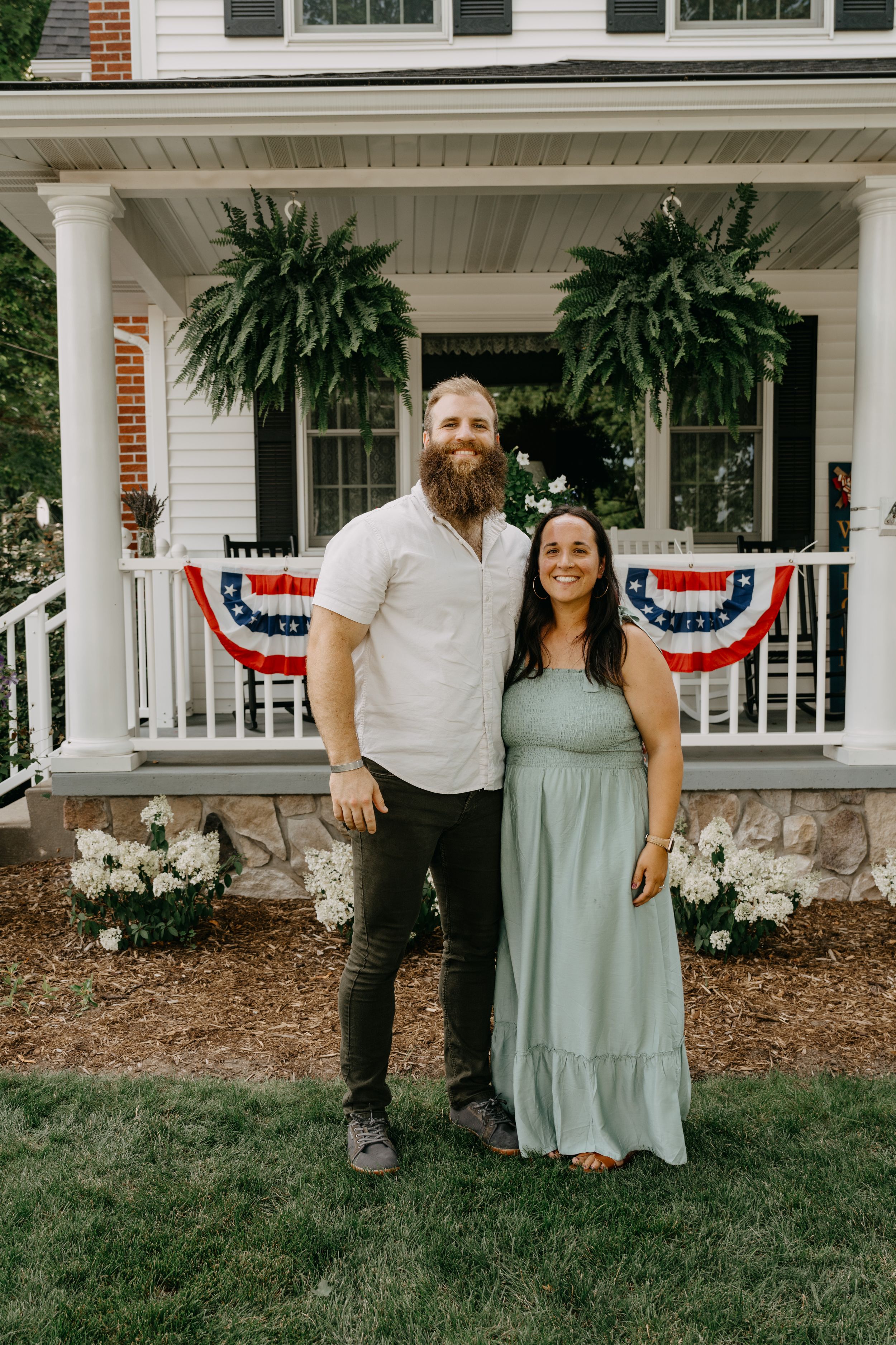 A couple stands together on their home's front porch adorned with patriotic decorations and lush hanging plants.