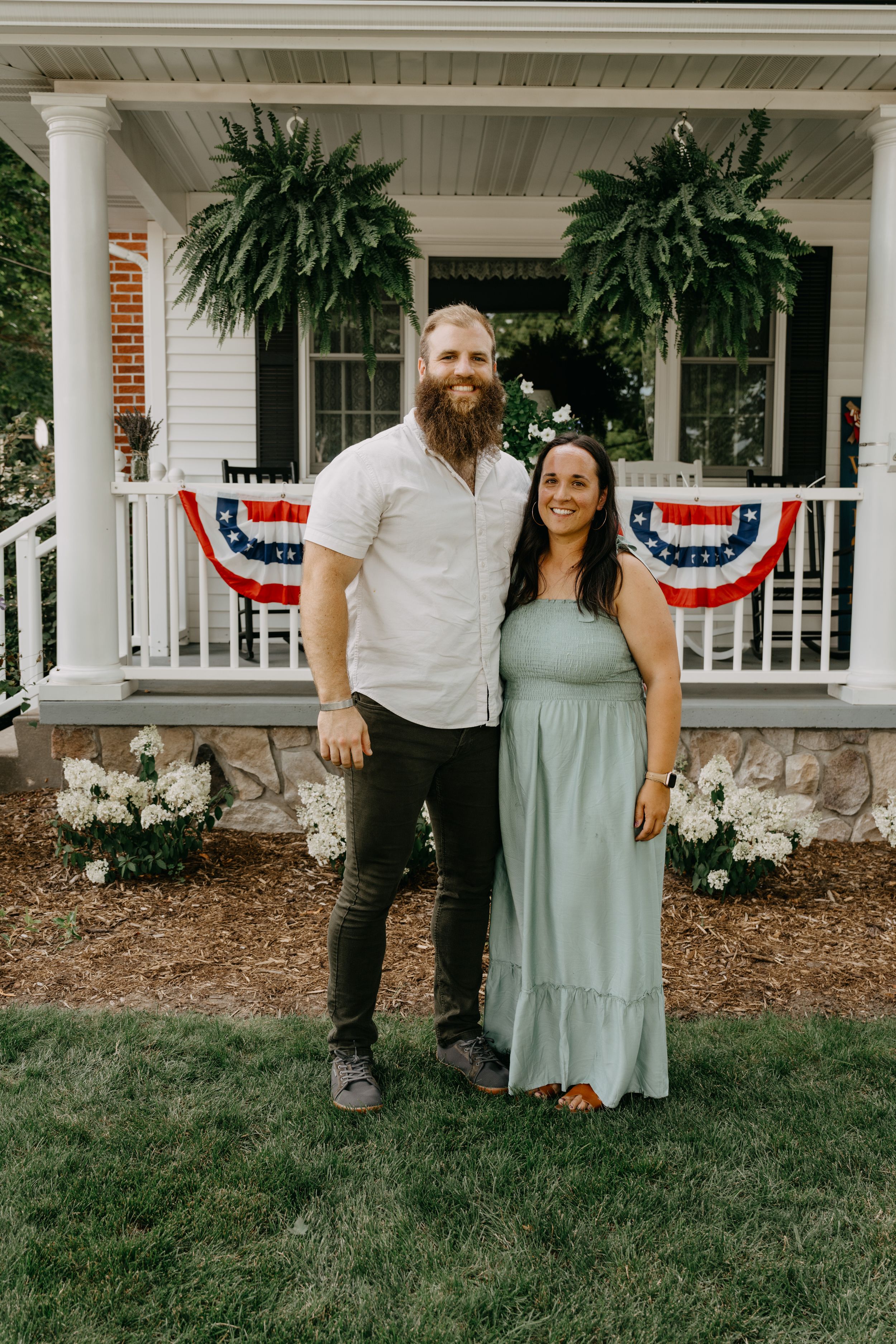 A couple poses in front of a white porch adorned with American flag bunting and lush ferns.