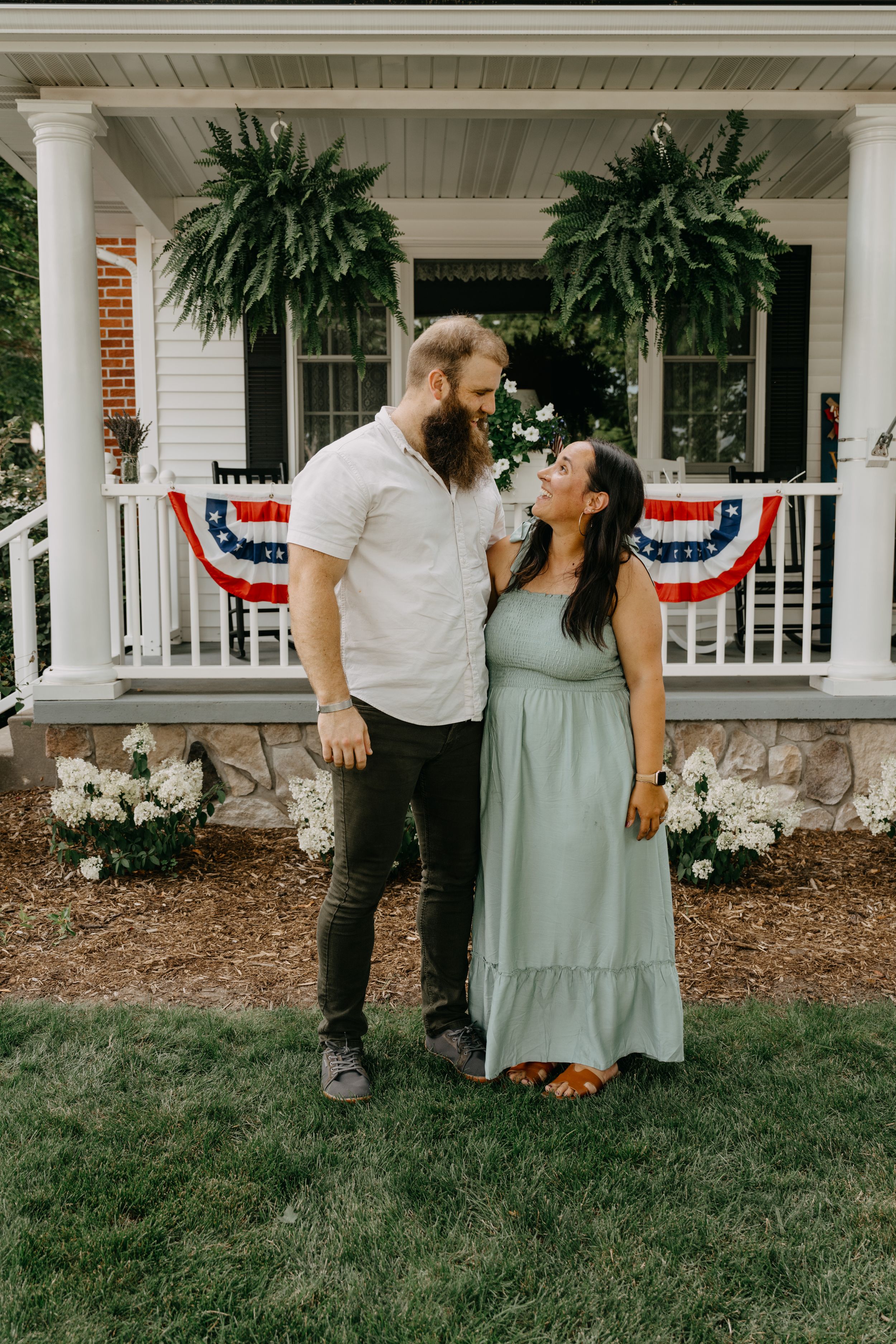A couple stands lovingly outside their home's front porch adorned with ferns and American flag bunting decor.