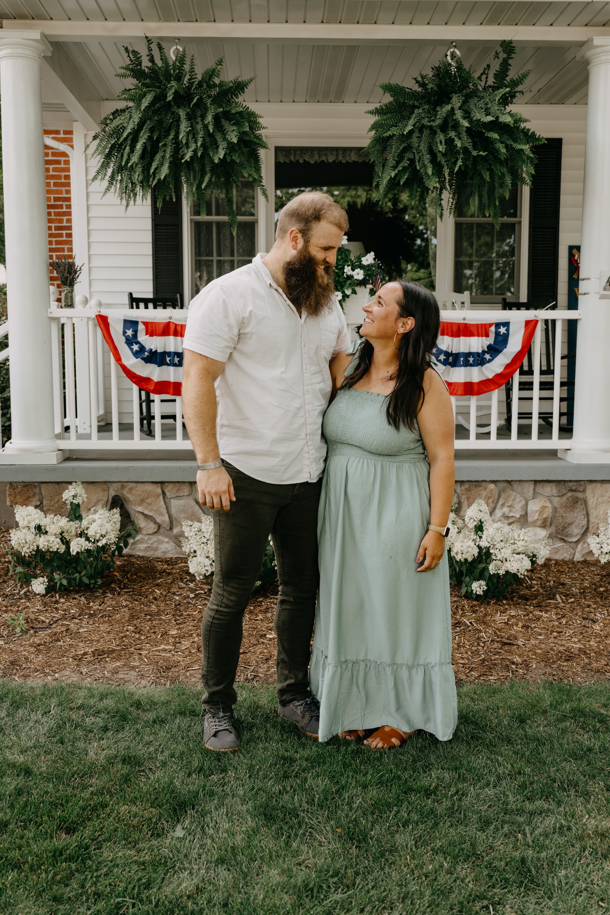 Couple stands together on a lawn in front of a porch decorated with patriotic bunting and large ferns.