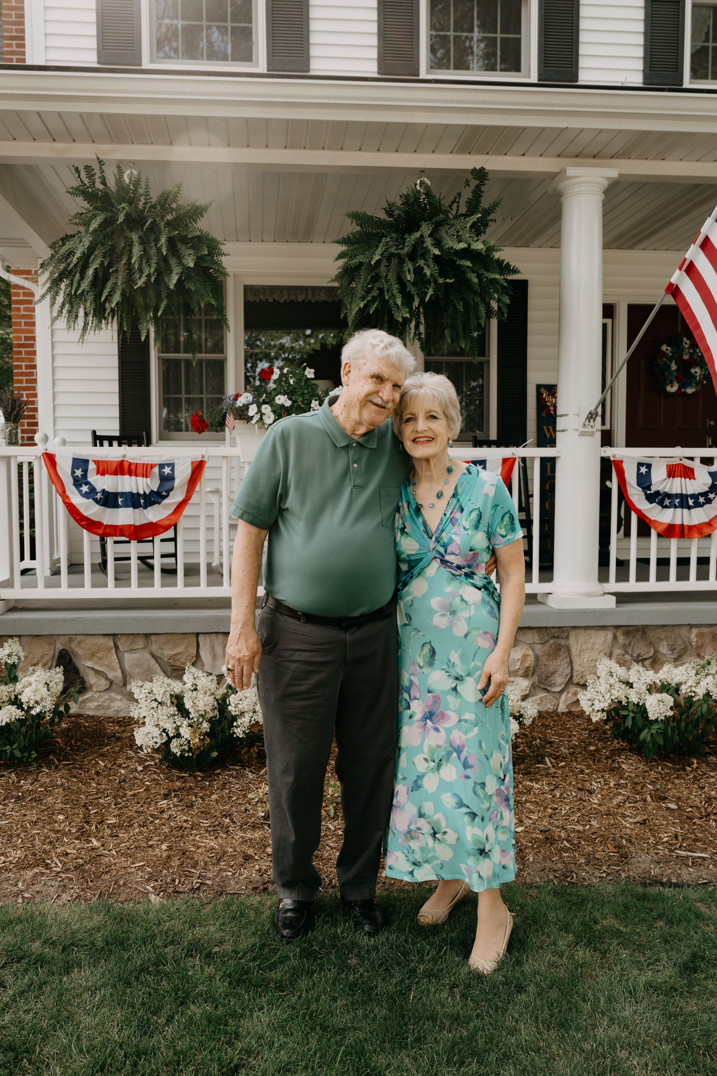 A couple stands together on a front porch decorated with American flag bunting and hanging ferns on a summer day.