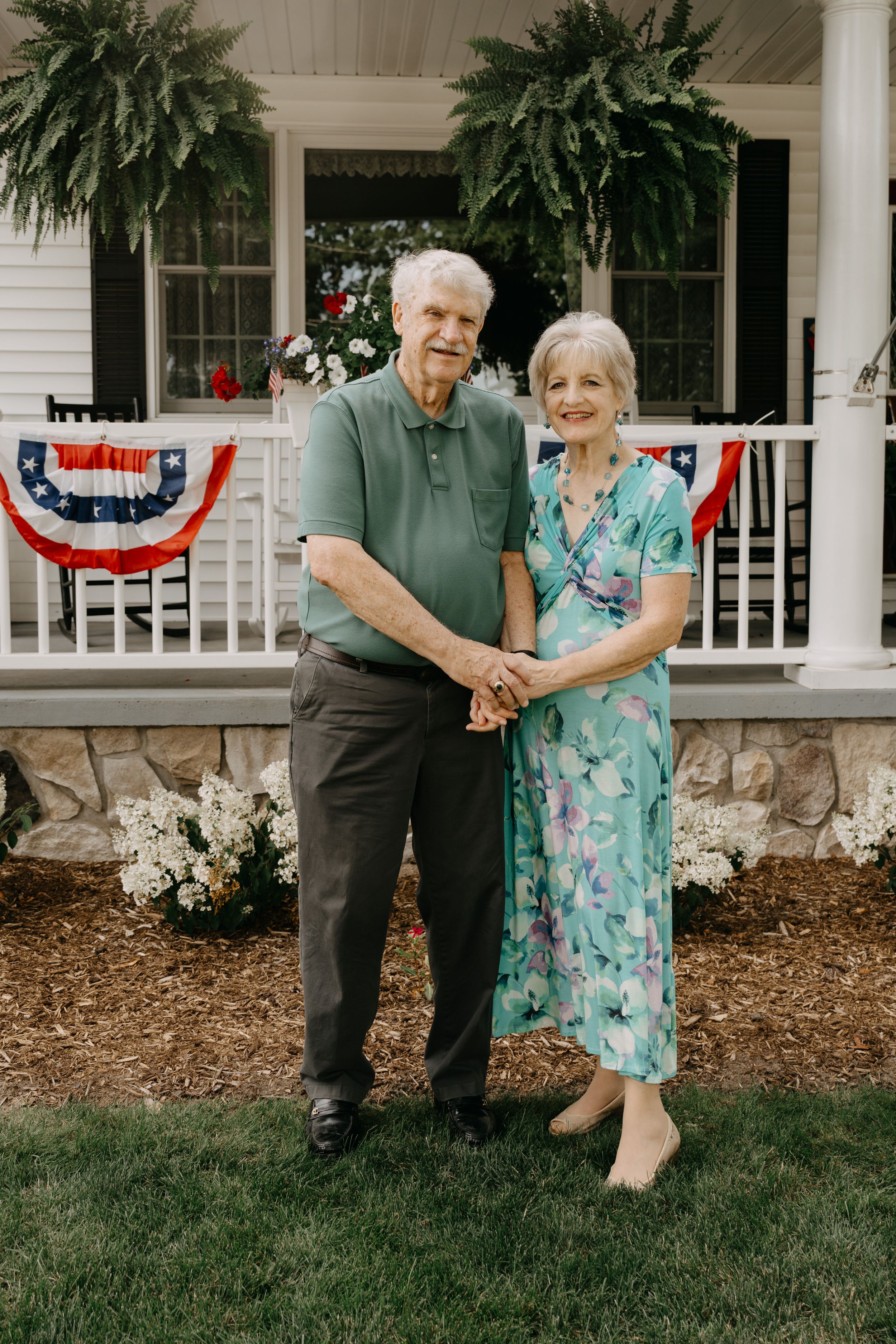 Elderly couple stands together on front porch with patriotic bunting and hanging ferns during summer.