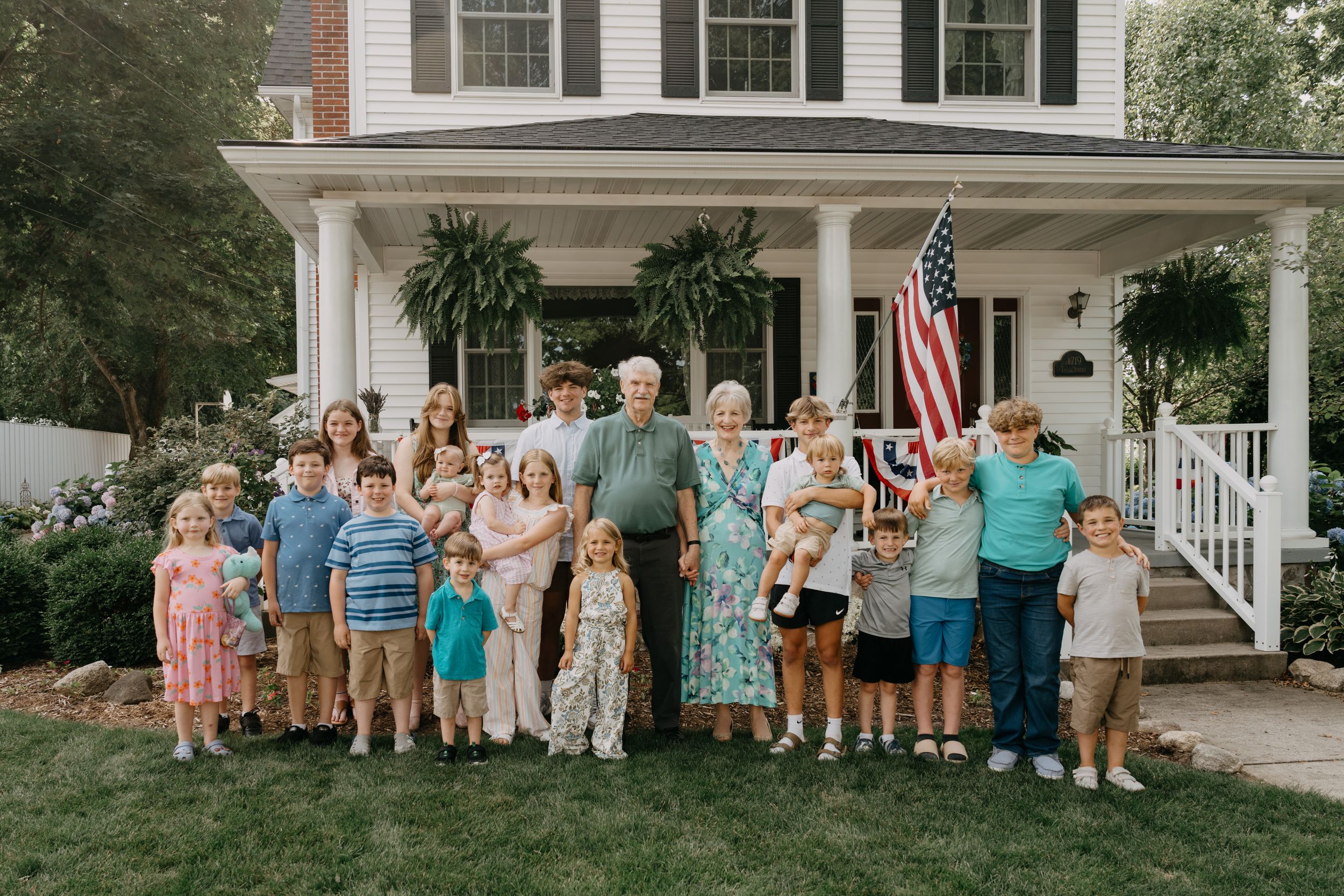A group of people gathered outdoors in front of a traditional white house with columns on a summer day.