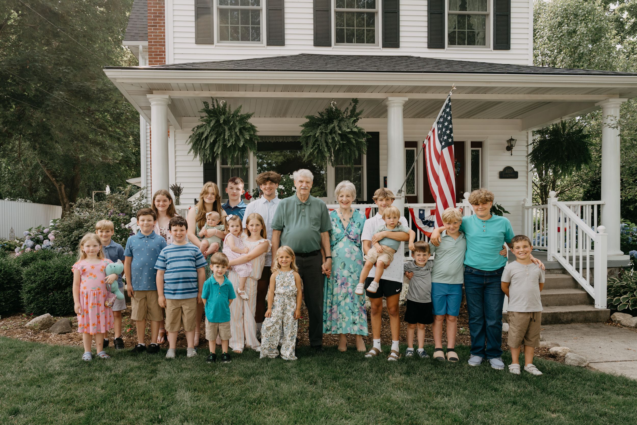 Large family group poses together on the front lawn of a traditional white colonial house with American flag and hanging plants.