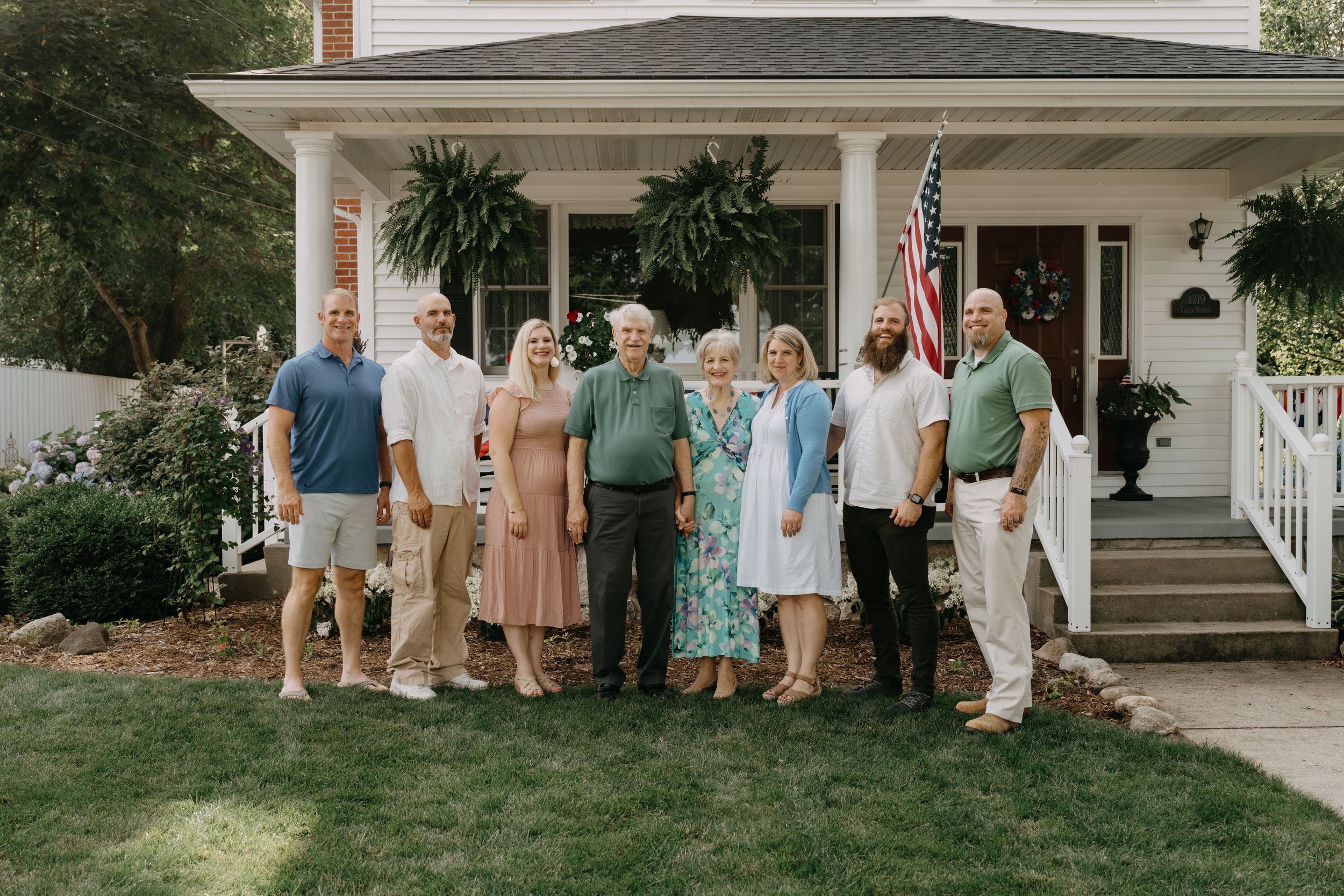 Group of people standing together on a front lawn in front of a white house with columns and an American flag.