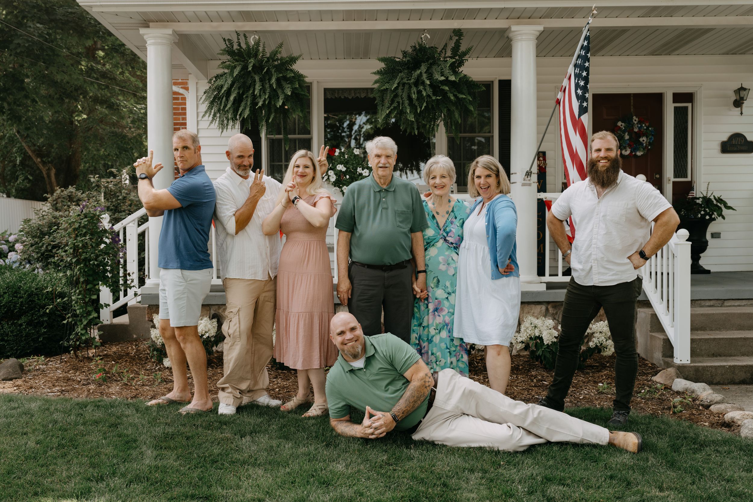 Group standing and posing casually on landscaped front porch of traditional home with American flag.
