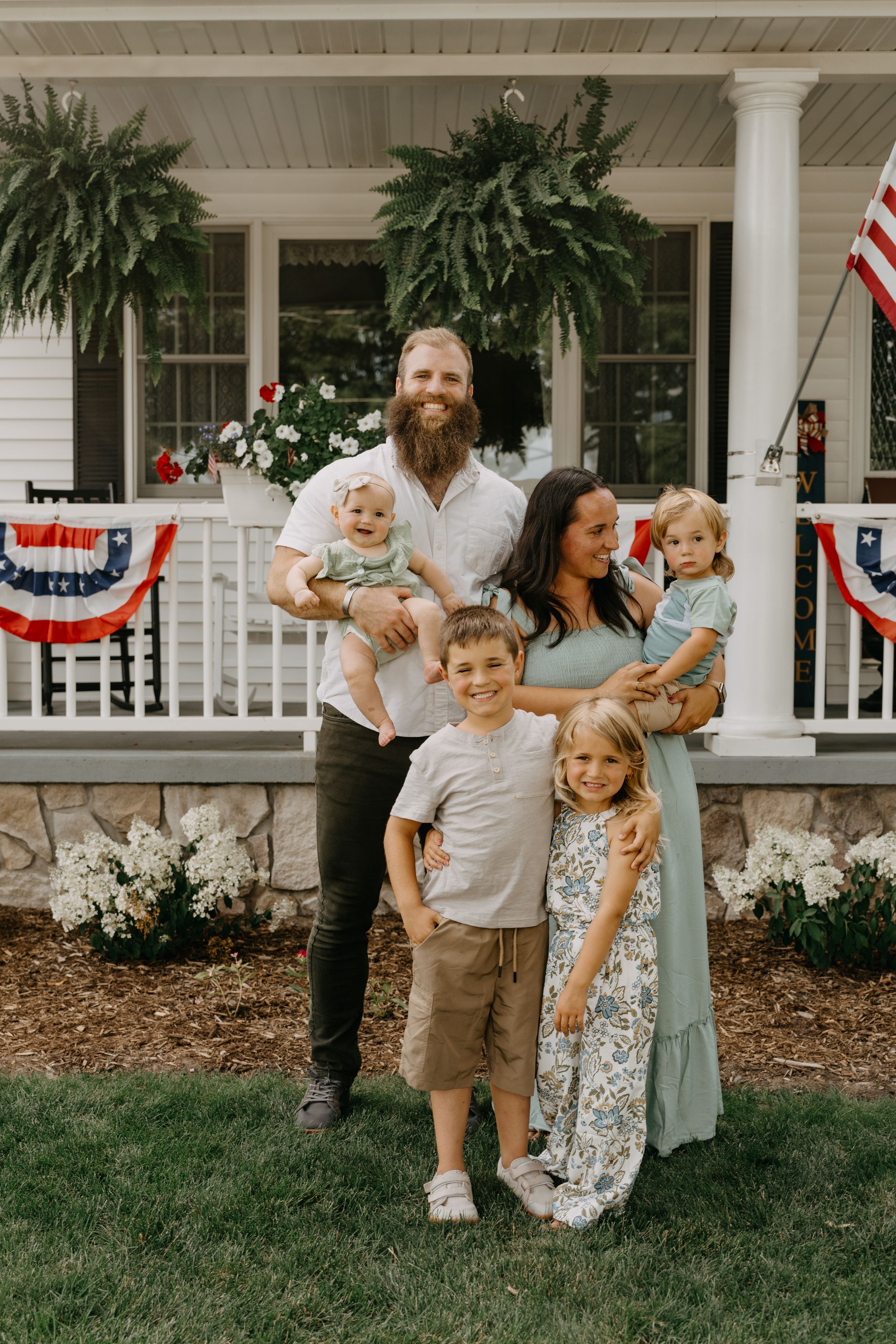 Family poses on front porch decorated with patriotic bunting and hanging ferns for Independence Day celebration.