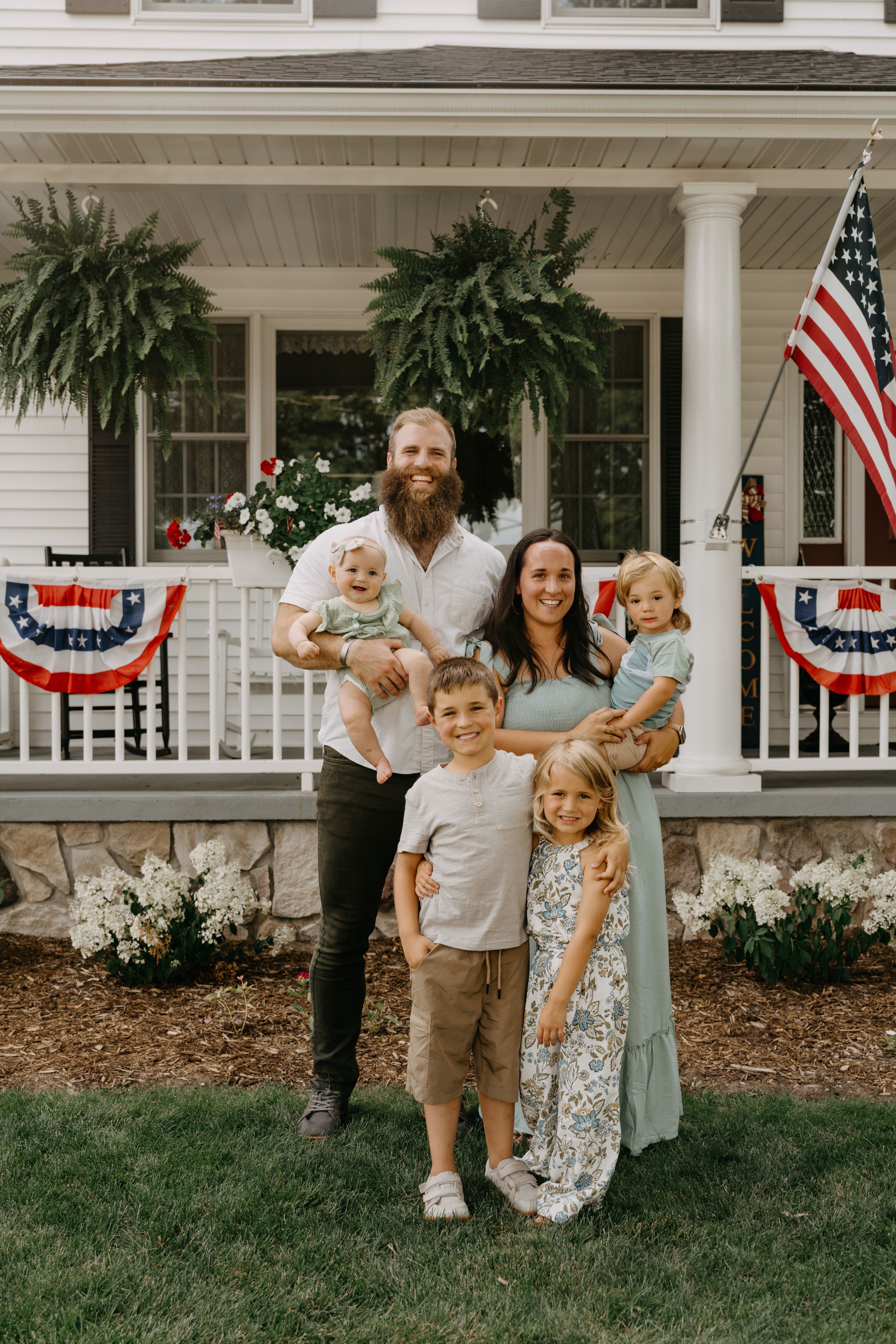 Patriotic family stands on front porch decorated with American flags and bunting during summer holiday celebration.