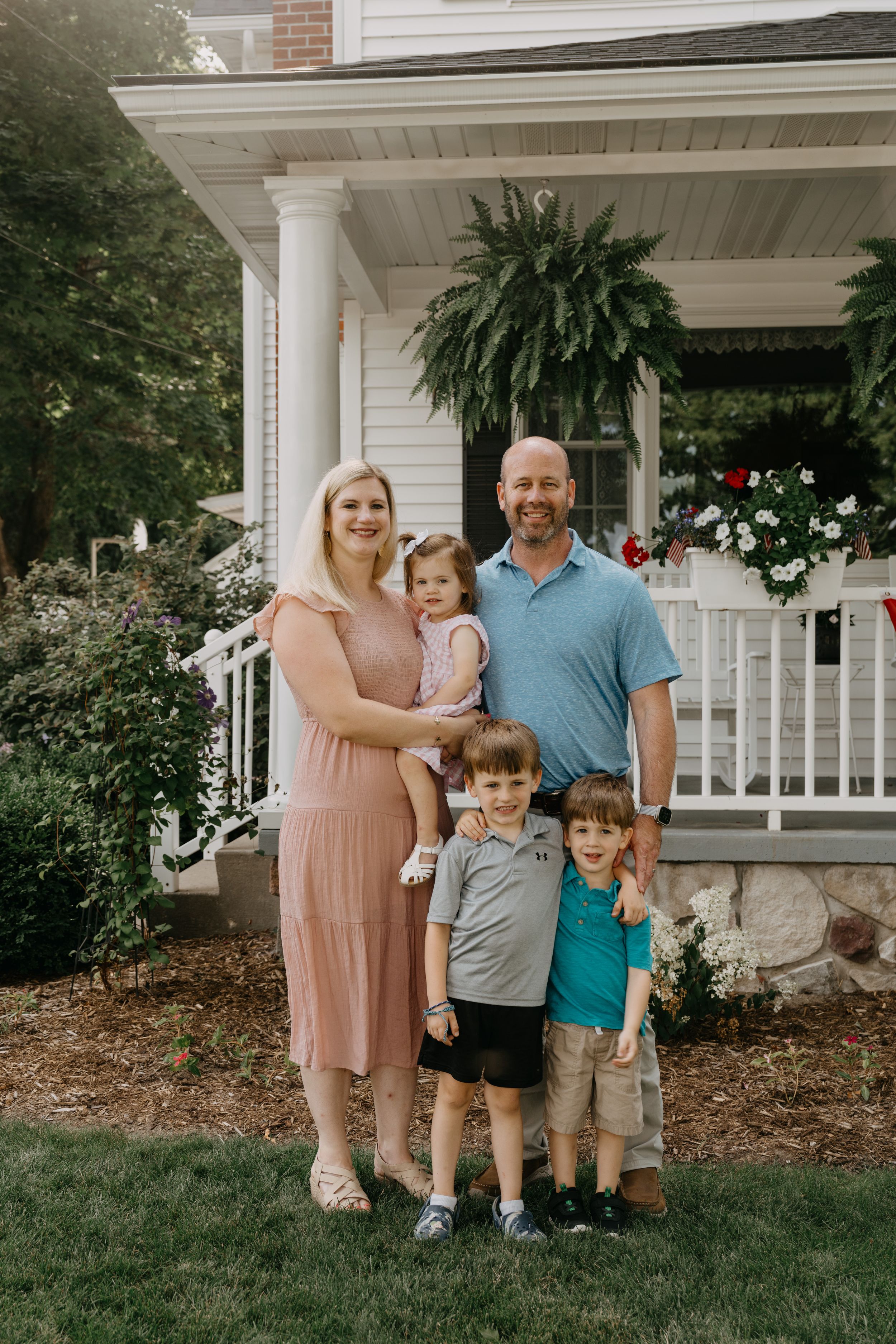 A family stands together on a green lawn in front of a white house with a covered porch decorated with hanging plants.