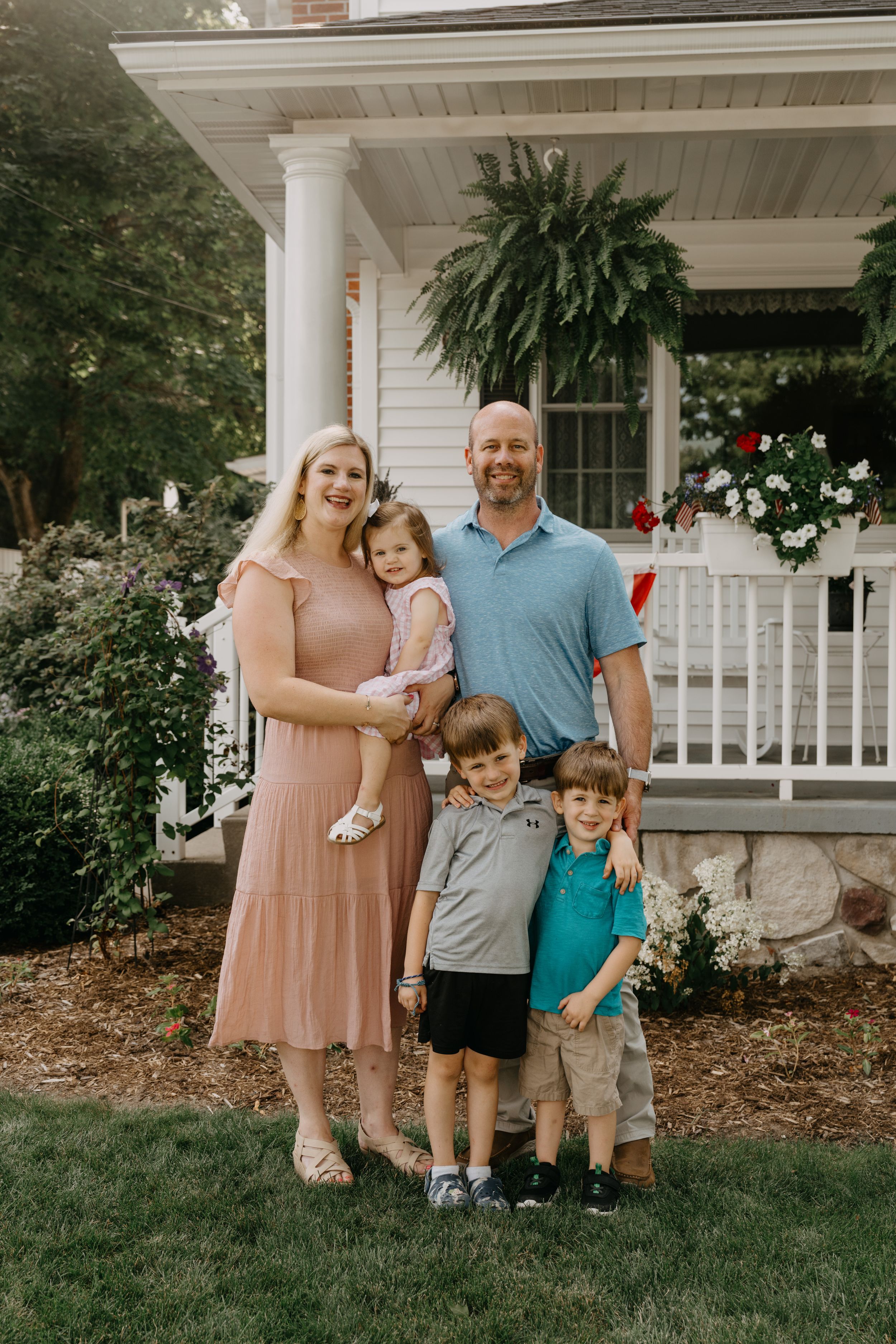 A loving family stands together on their front porch with white columns and hanging ferns on a summer day.
