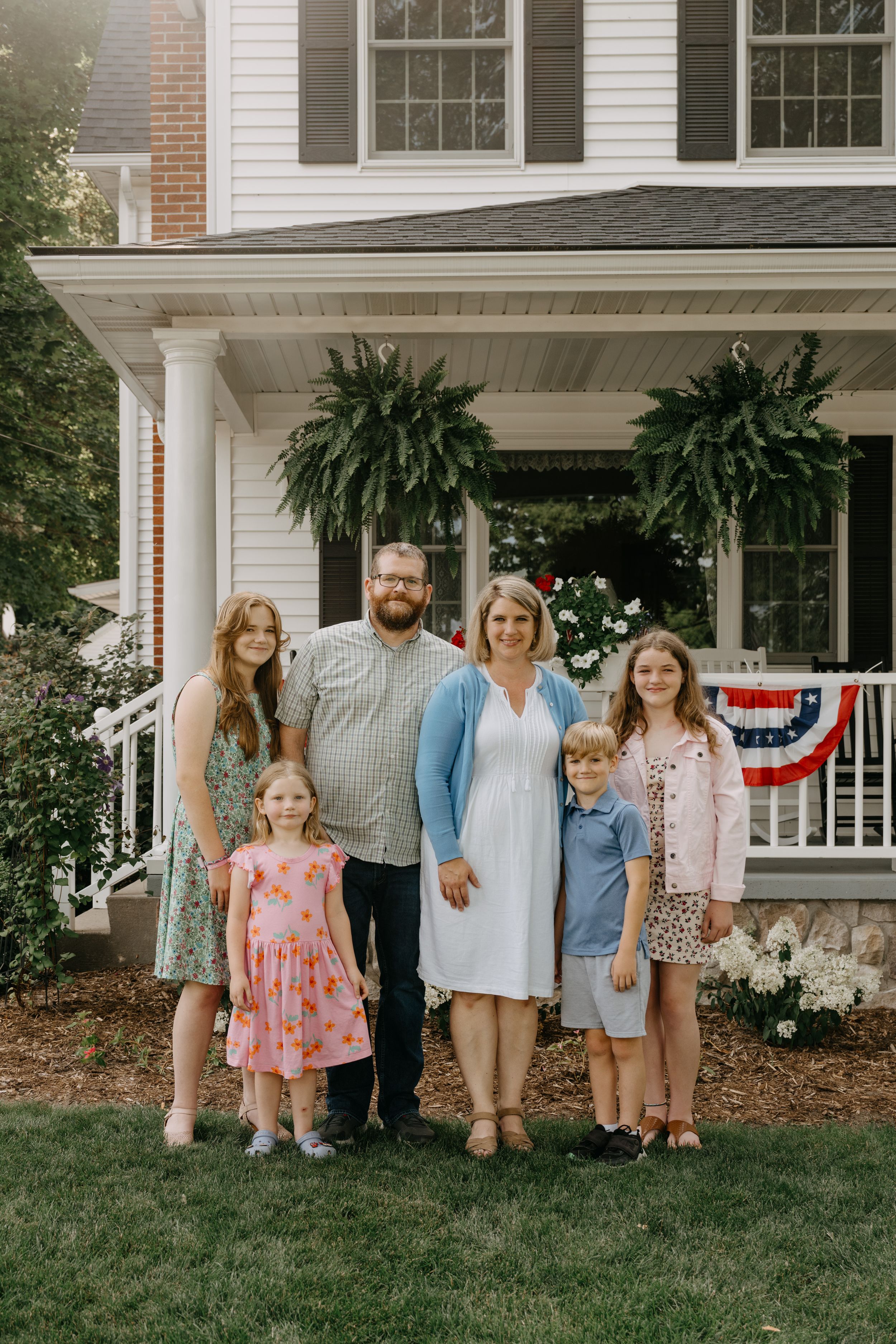 A family poses together on their lawn in front of a white colonial house with a patriotic bunting-decorated porch.