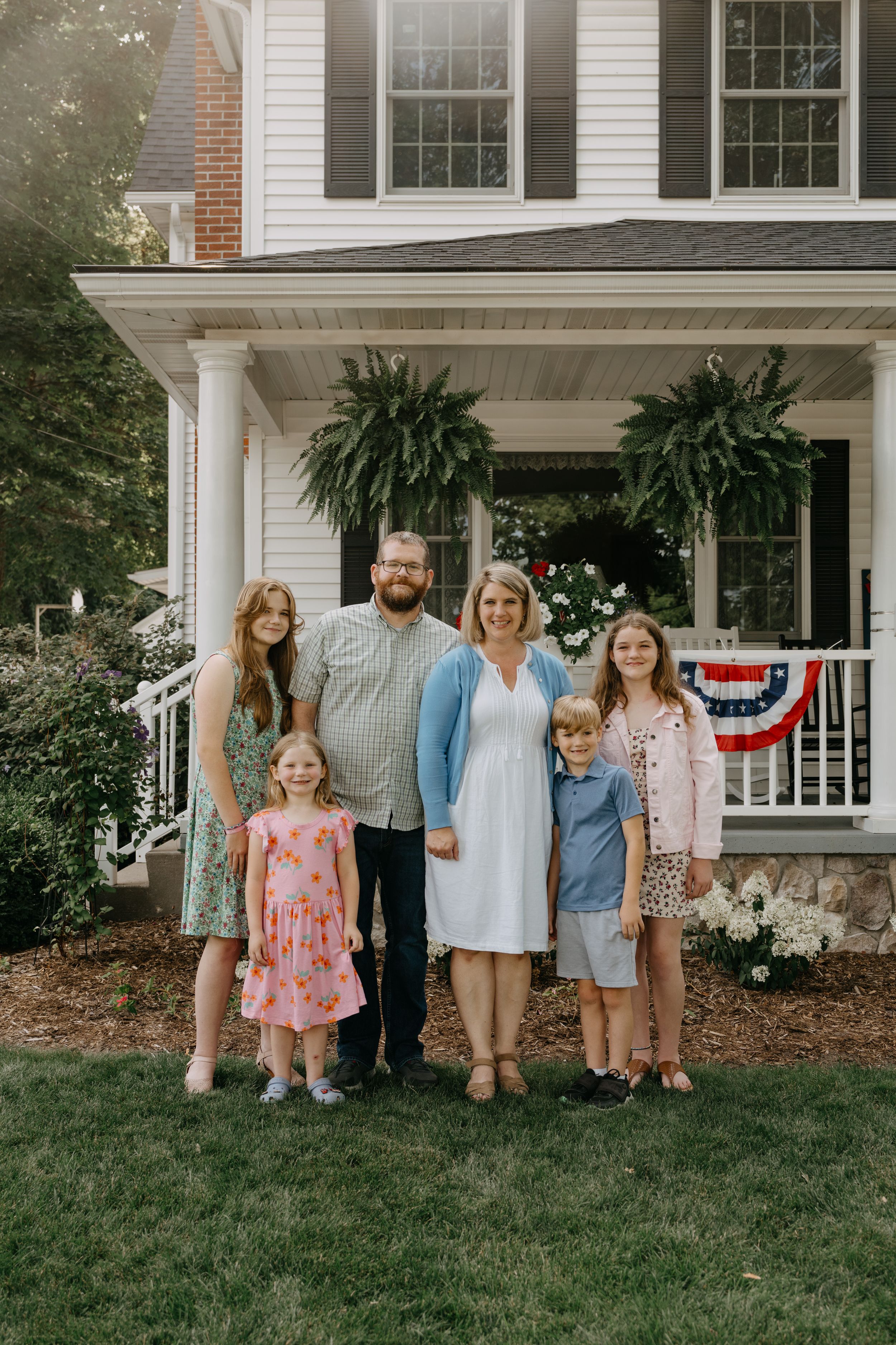 A family stands on their front lawn in front of a white colonial house with large hanging ferns and patriotic bunting.