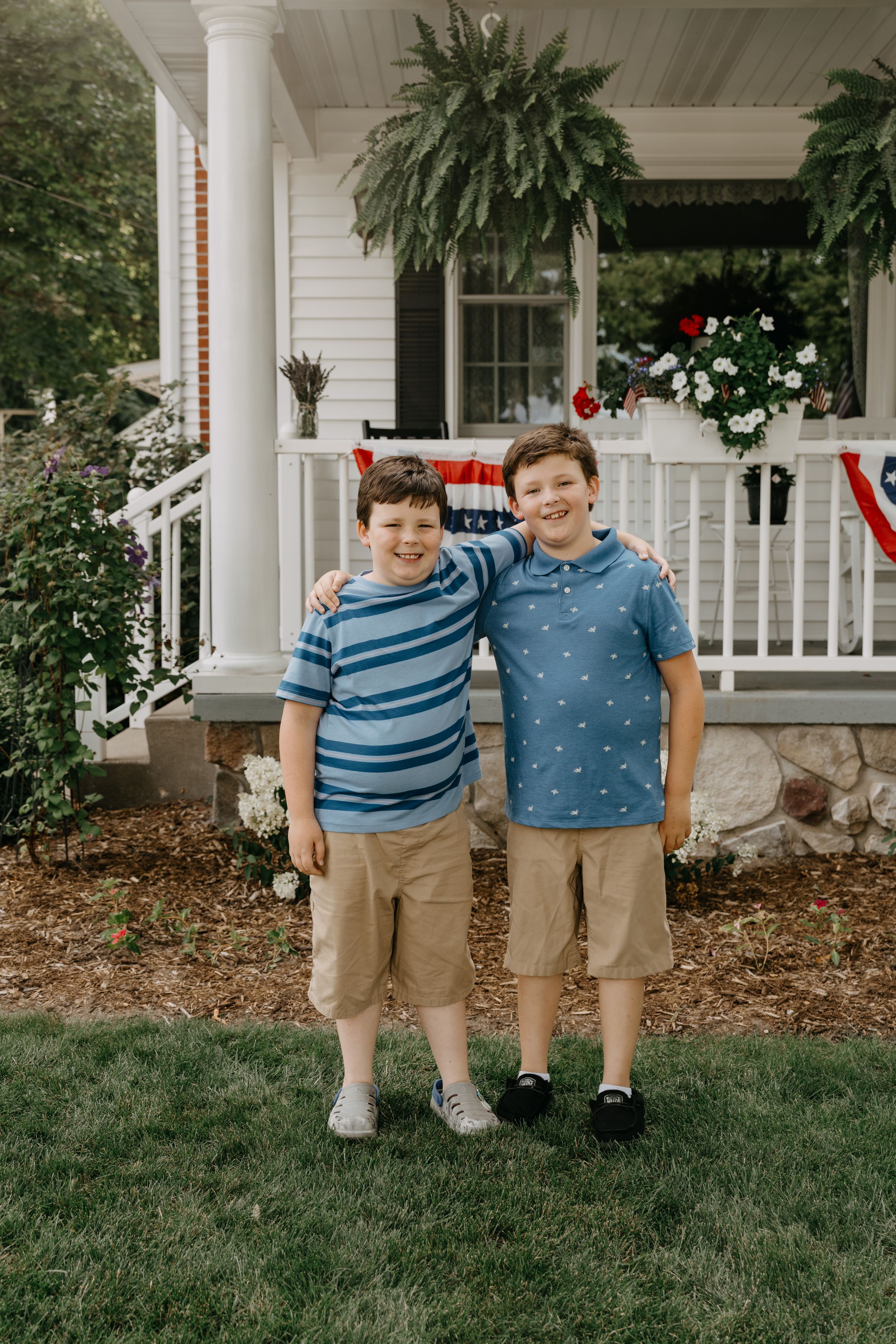 Two young figures in casual attire stand on grass in front of a white porch decorated with hanging plants and patriotic bunting.
