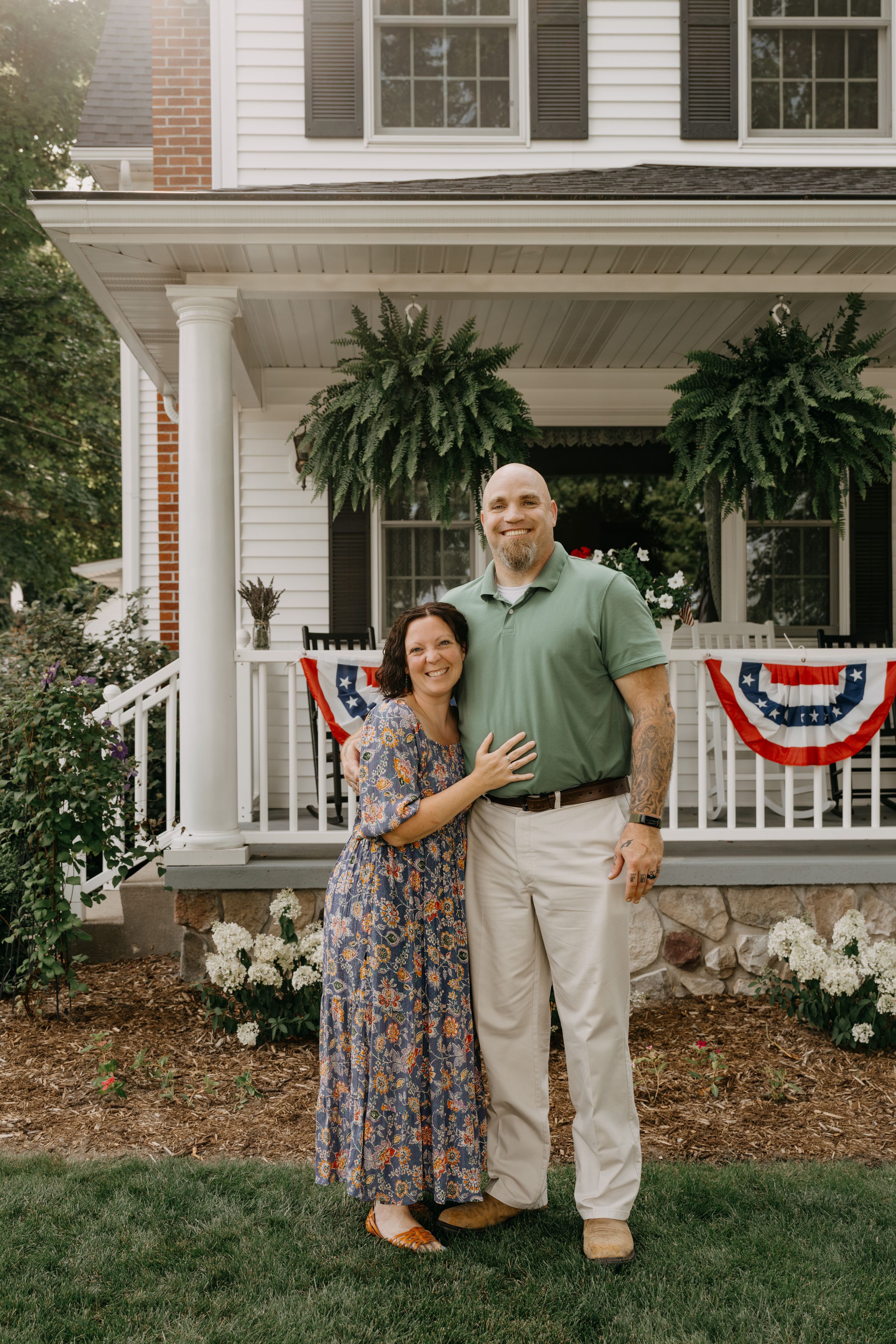 A couple stands on their patriotic porch decorated with American flag buntings and hanging ferns on a summer day.