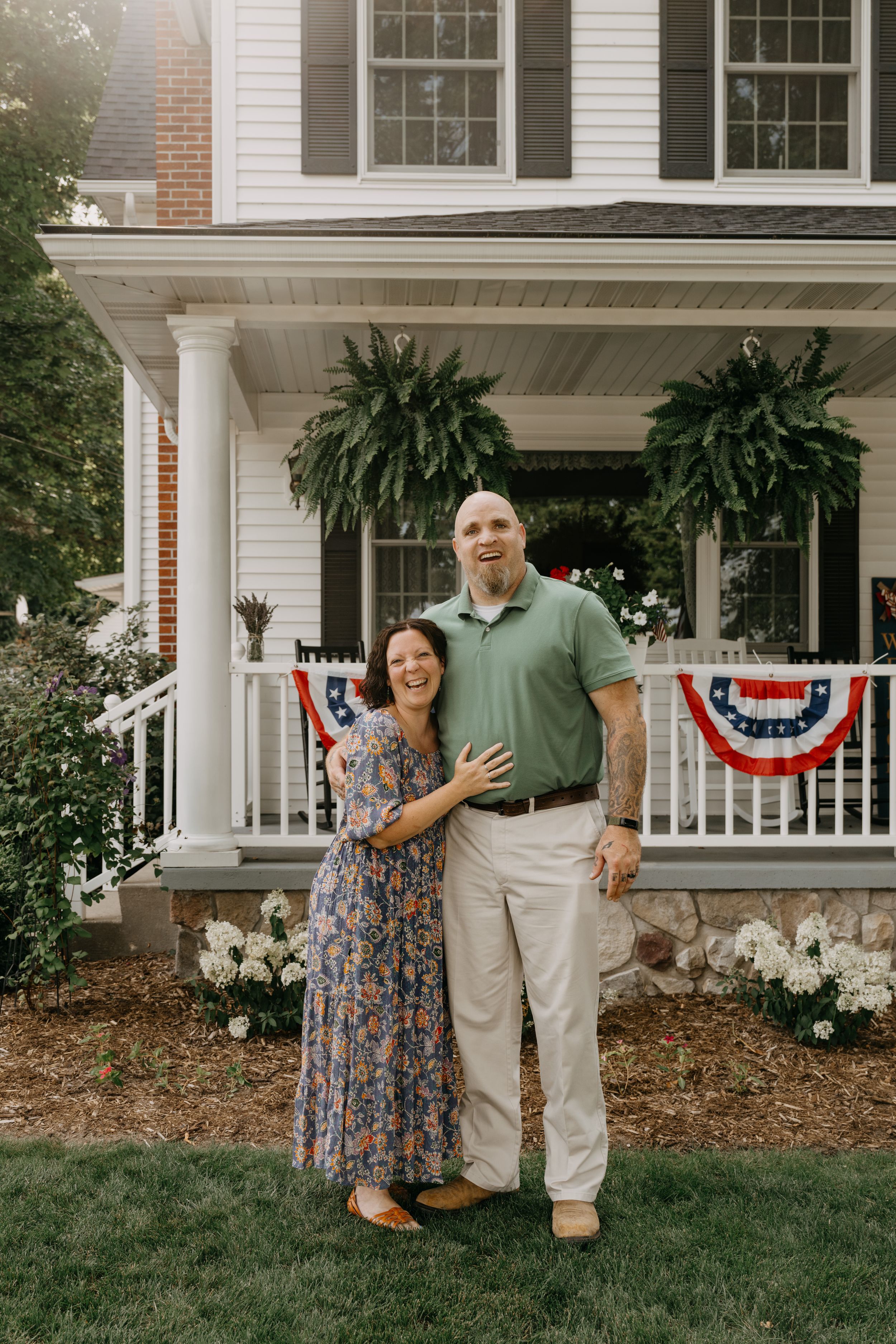 A couple poses together on their classic white porch decorated with patriotic bunting and hanging ferns.