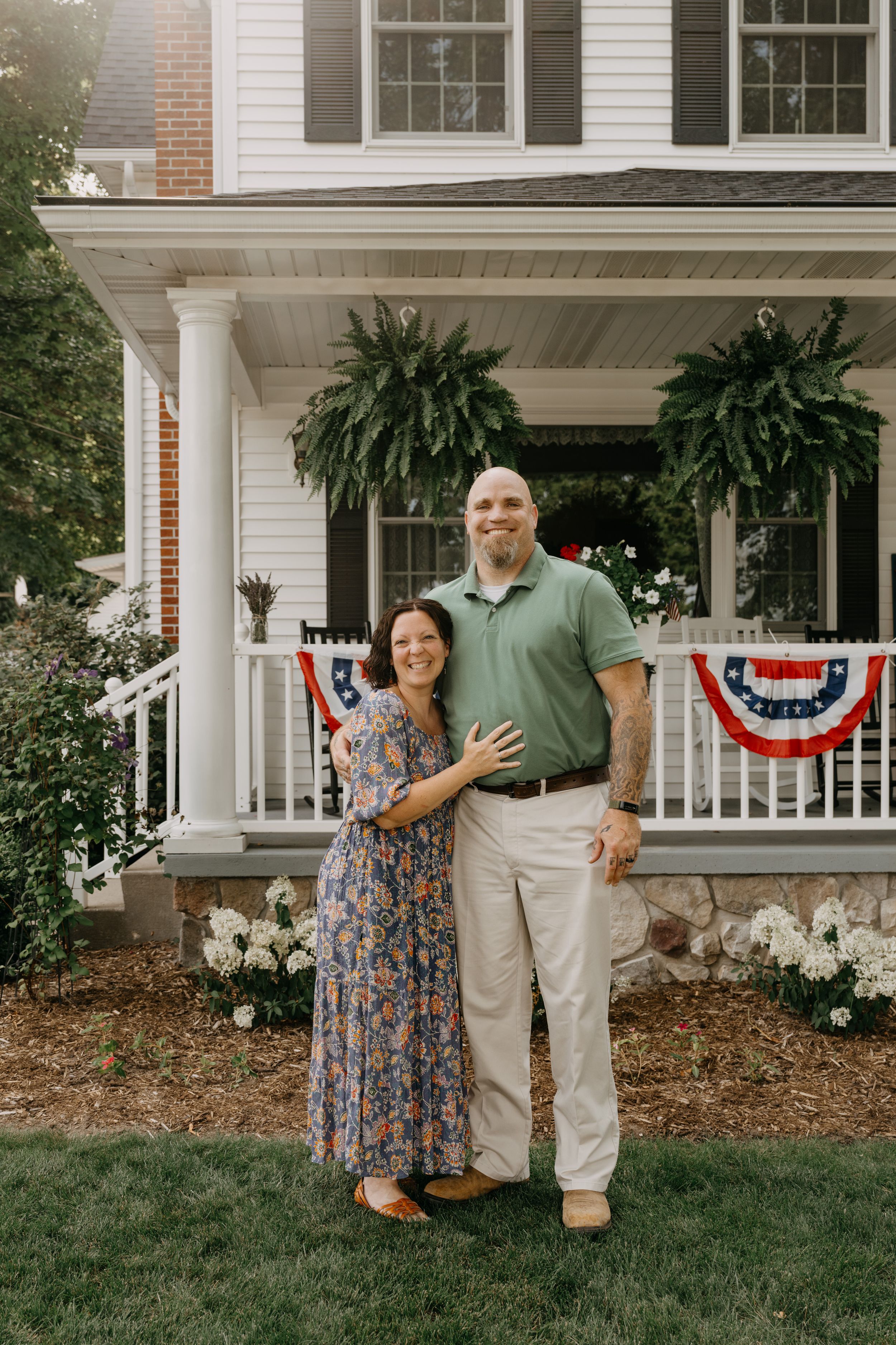 A couple stands together on their lawn in front of a classic white porch decorated with patriotic bunting and ferns.