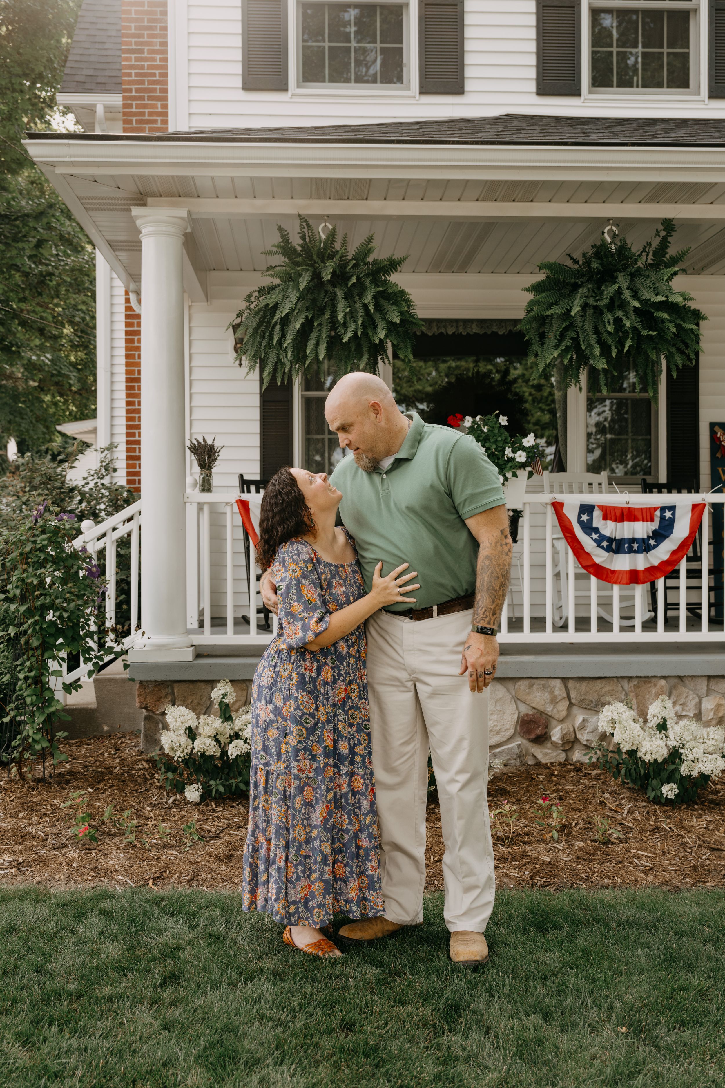 A couple shares an intimate moment together on their front lawn with patriotic decor and hanging ferns on the porch.