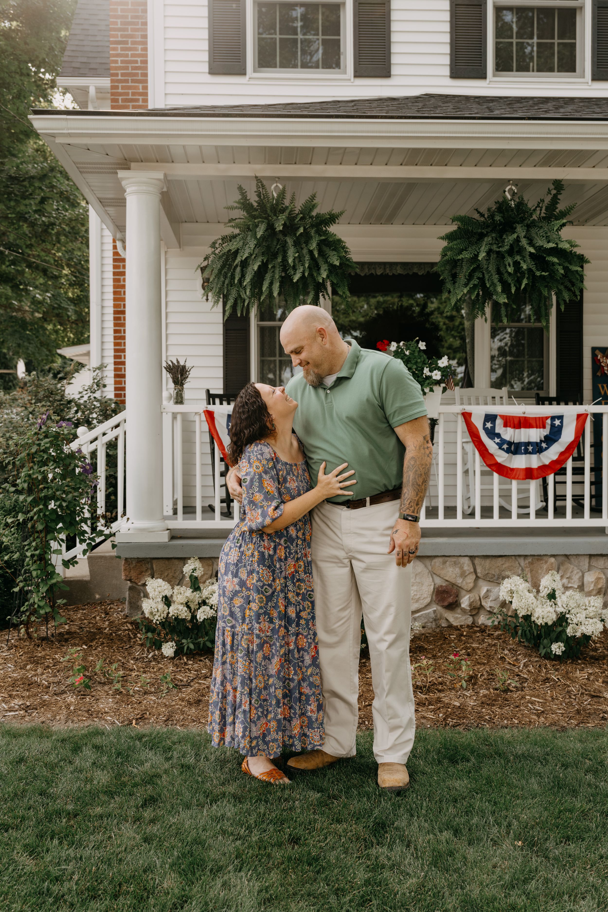 Two people standing together on the lawn in front of a white house with patriotic bunting on the porch railings.