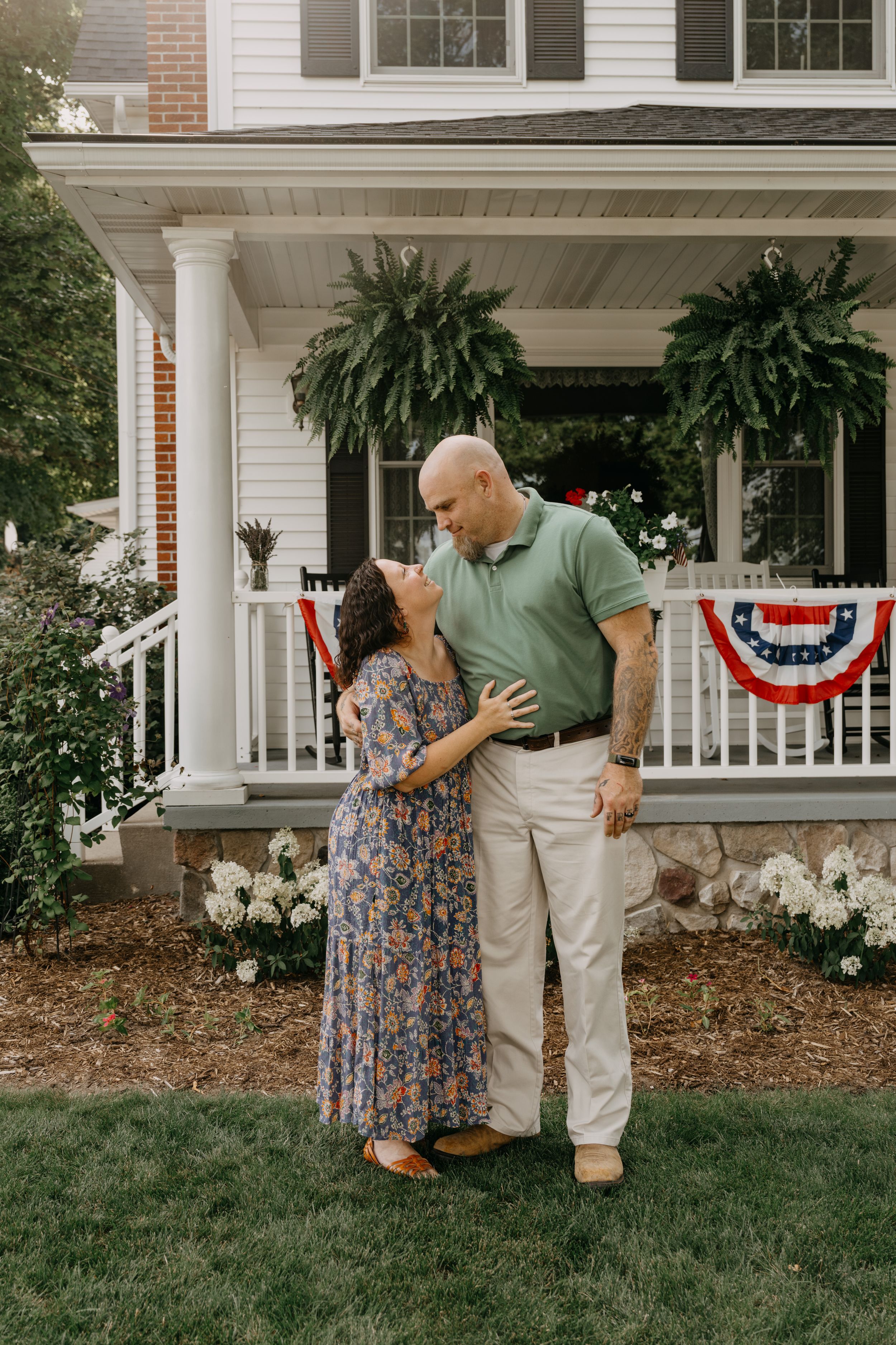 A couple embraces on a porch decorated with hanging ferns and patriotic bunting in front of a traditional white house.