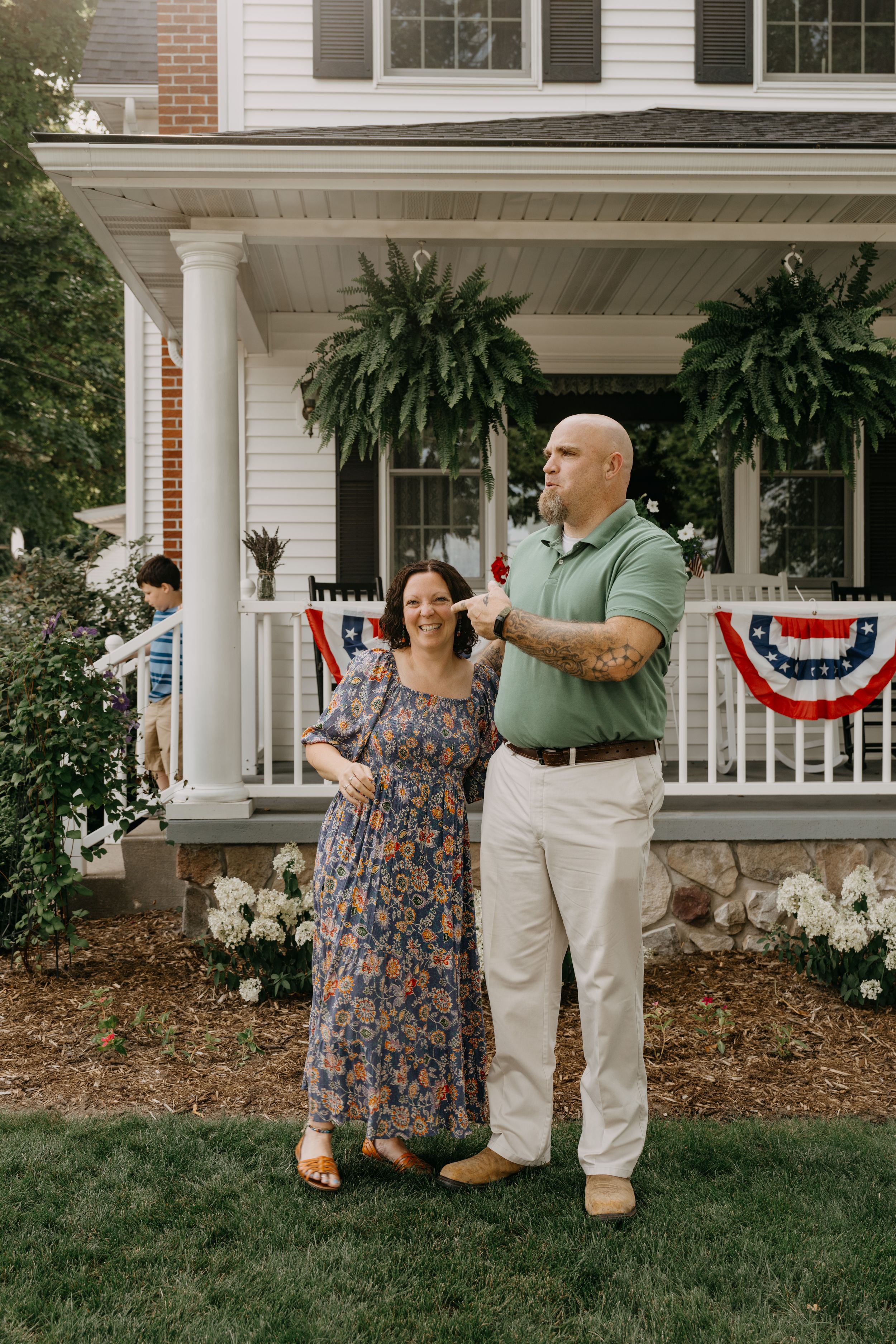 Two people pose on the lawn in front of a white house with patriotic bunting and hanging ferns on the porch.