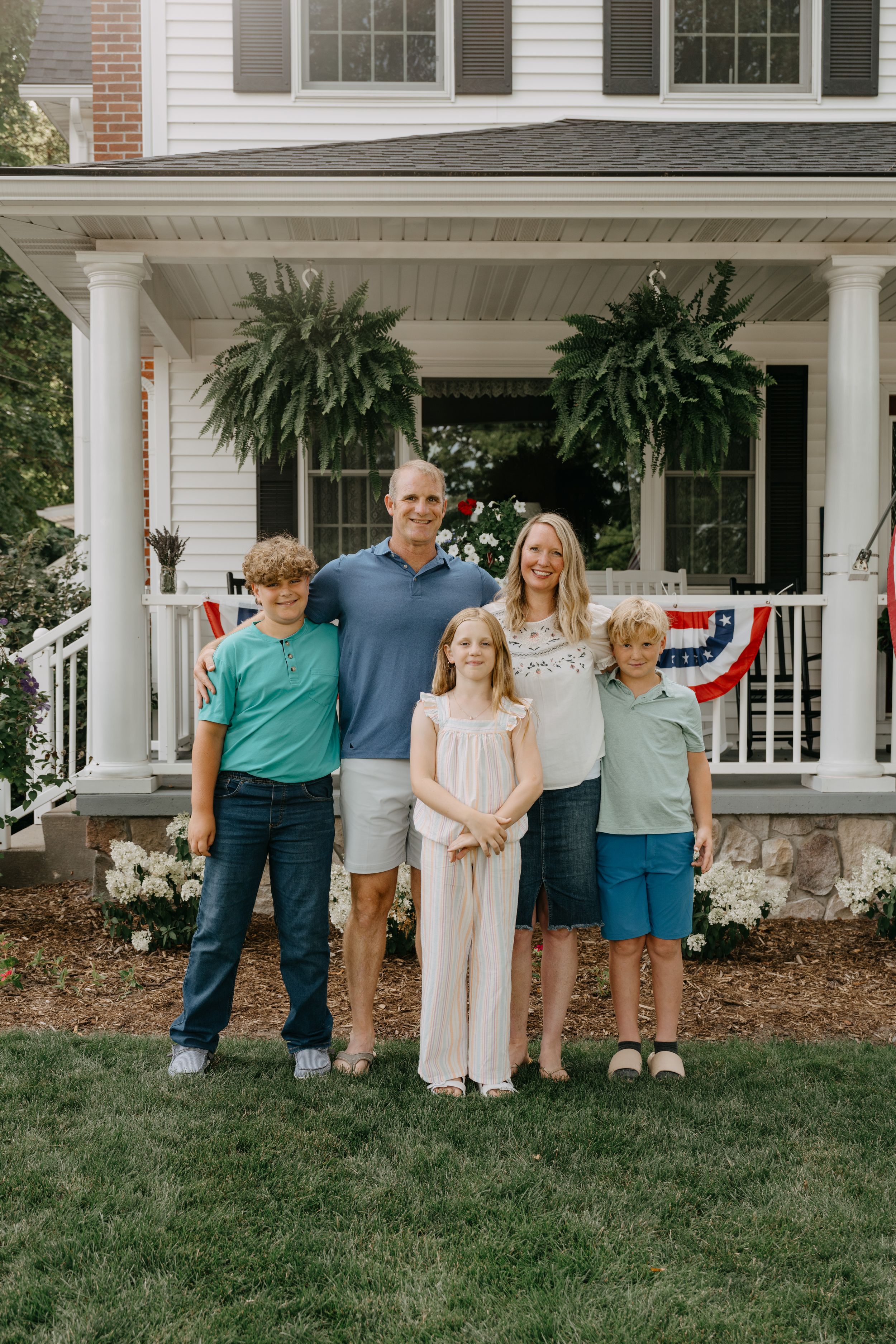 A family of five stands together on their front lawn against a classic American porch with patriotic decorations.