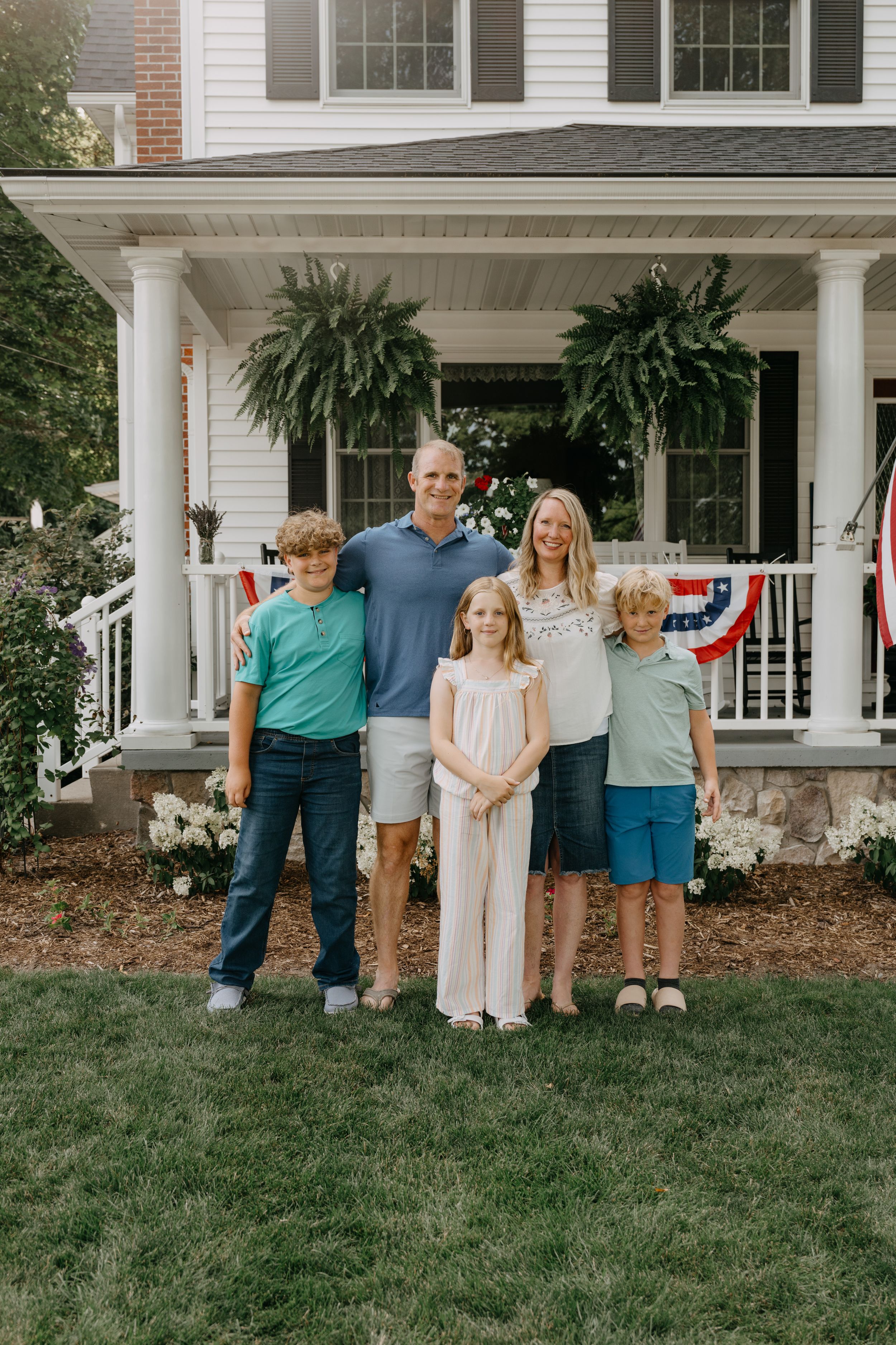 Family stands together on a colonial style front porch with white columns and hanging ferns, decorated with patriotic bunting.