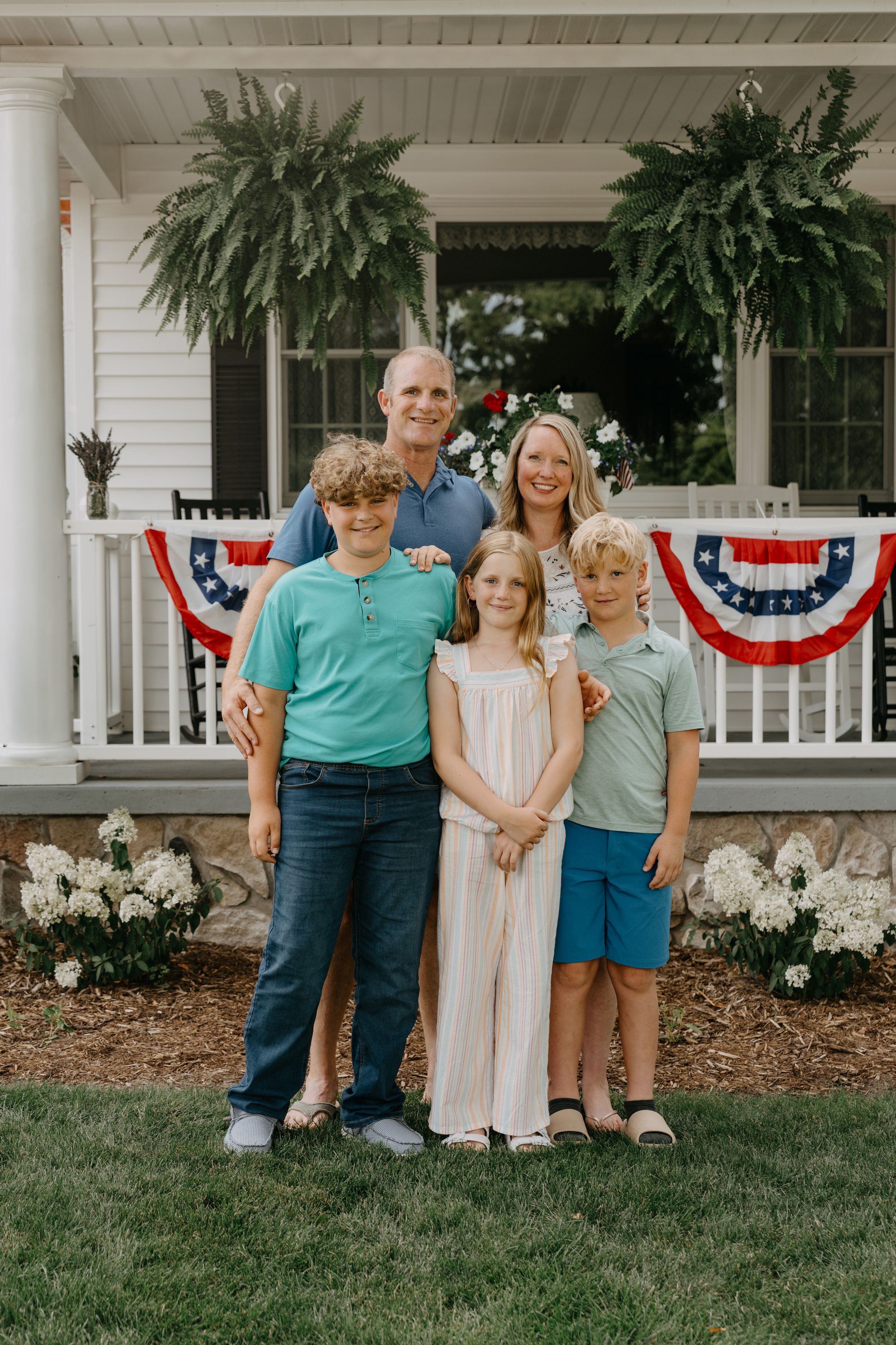 A family poses on a porch decorated with patriotic bunting and hanging ferns on a summer day.