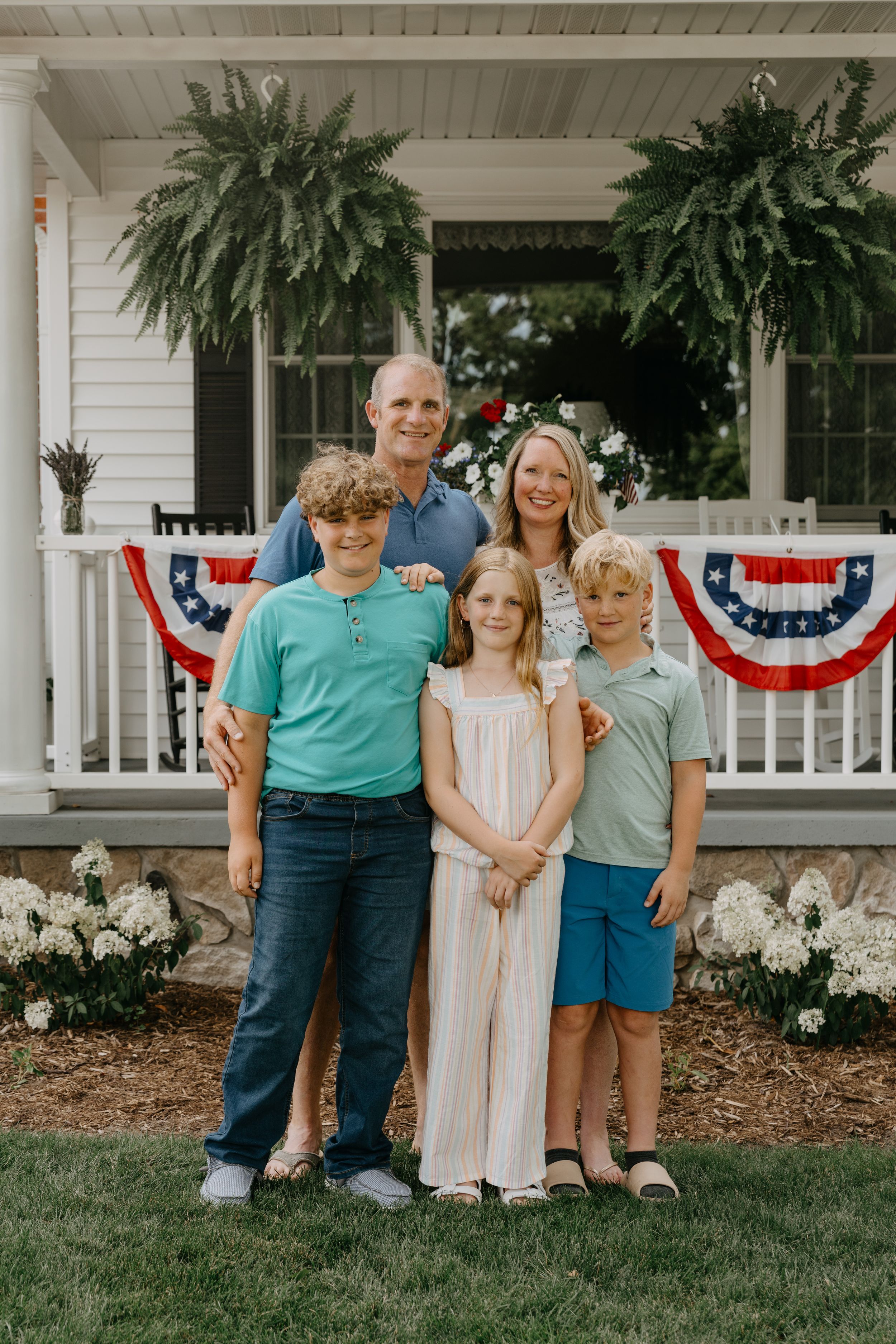 A family group poses together on a front porch adorned with American flag buntings and hanging ferns.