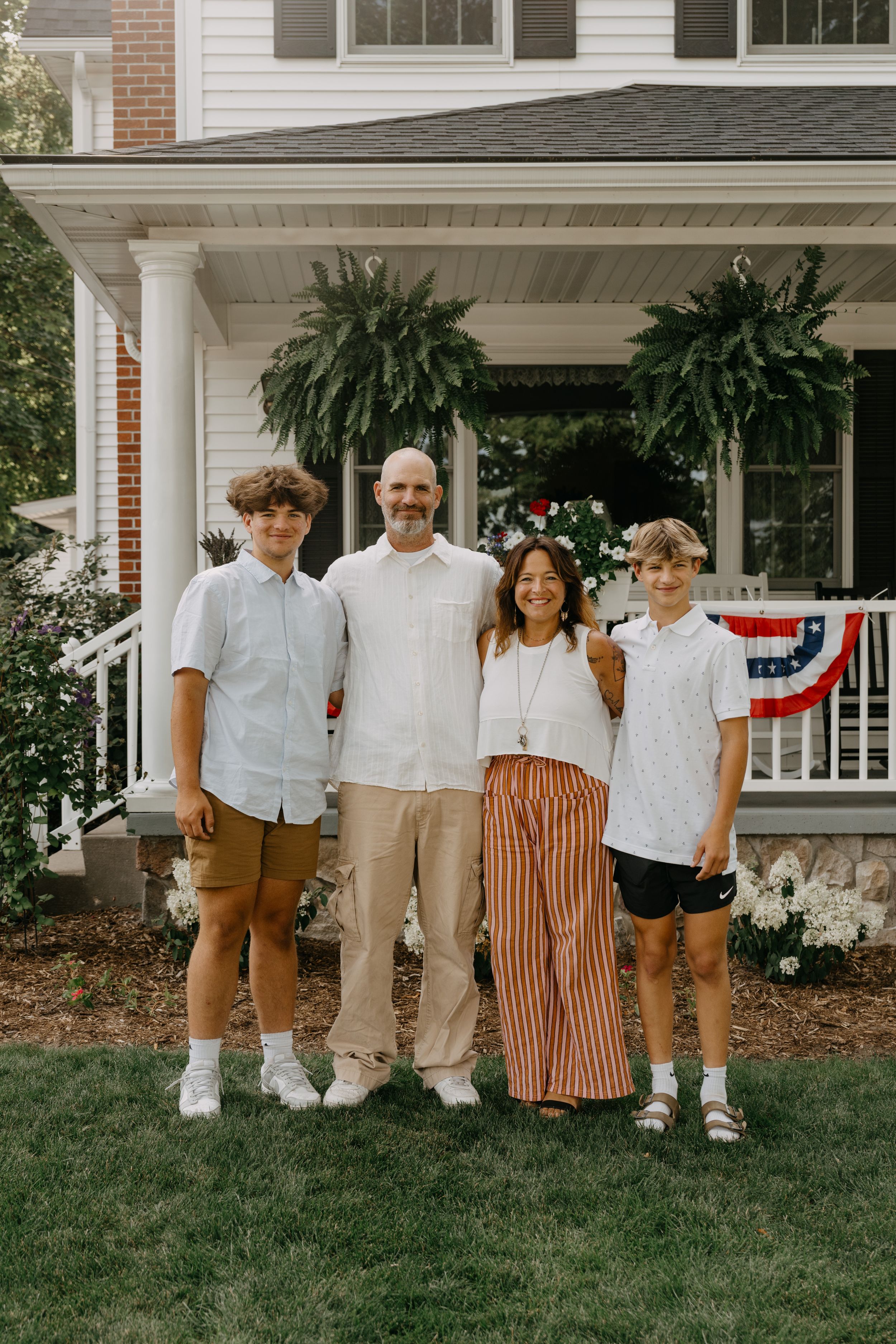 Four people stand together on a lawn in front of a white house with patriotic bunting and ferns on the porch.