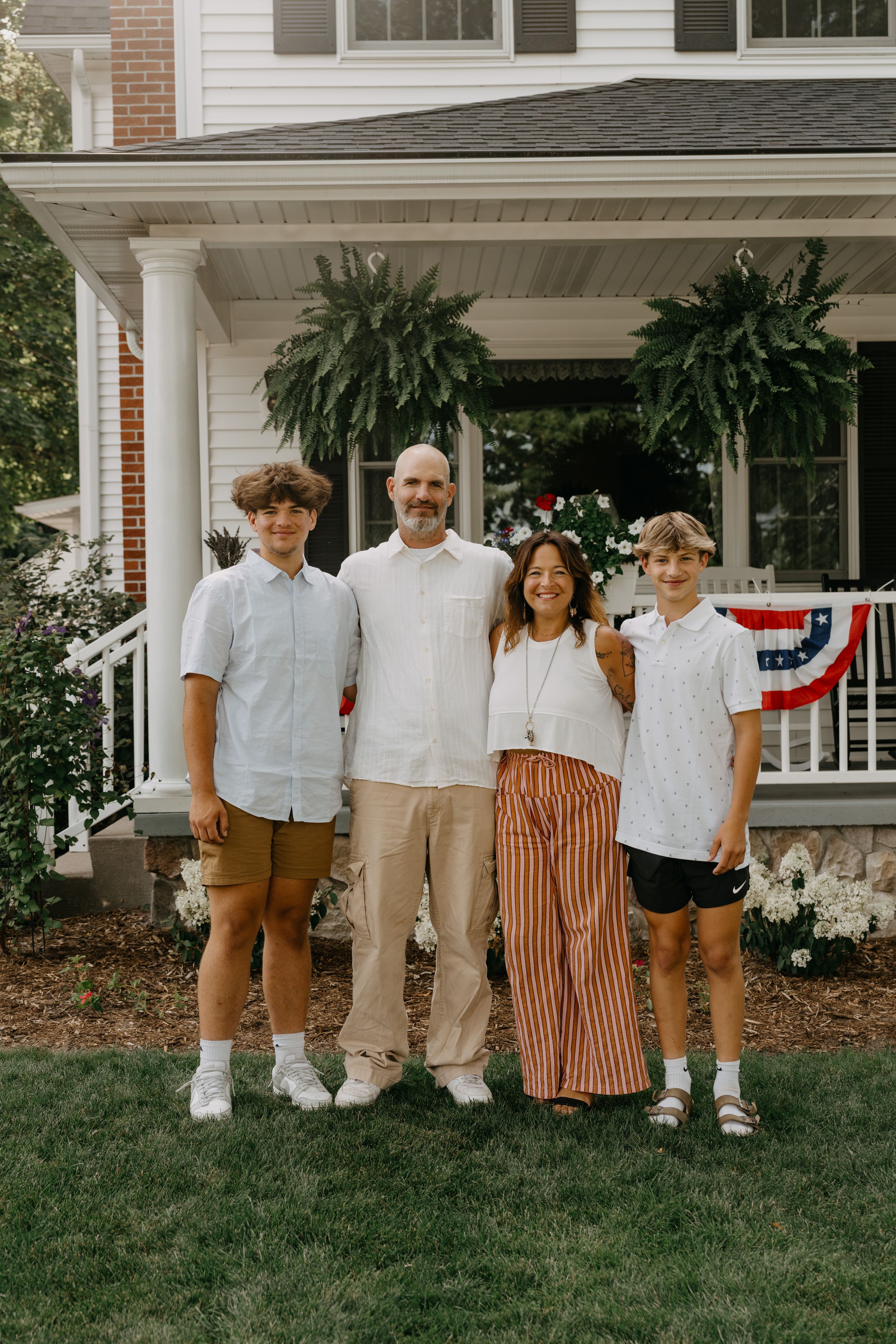 A family stands together on their front lawn in front of a porch decorated with hanging ferns and patriotic bunting.
