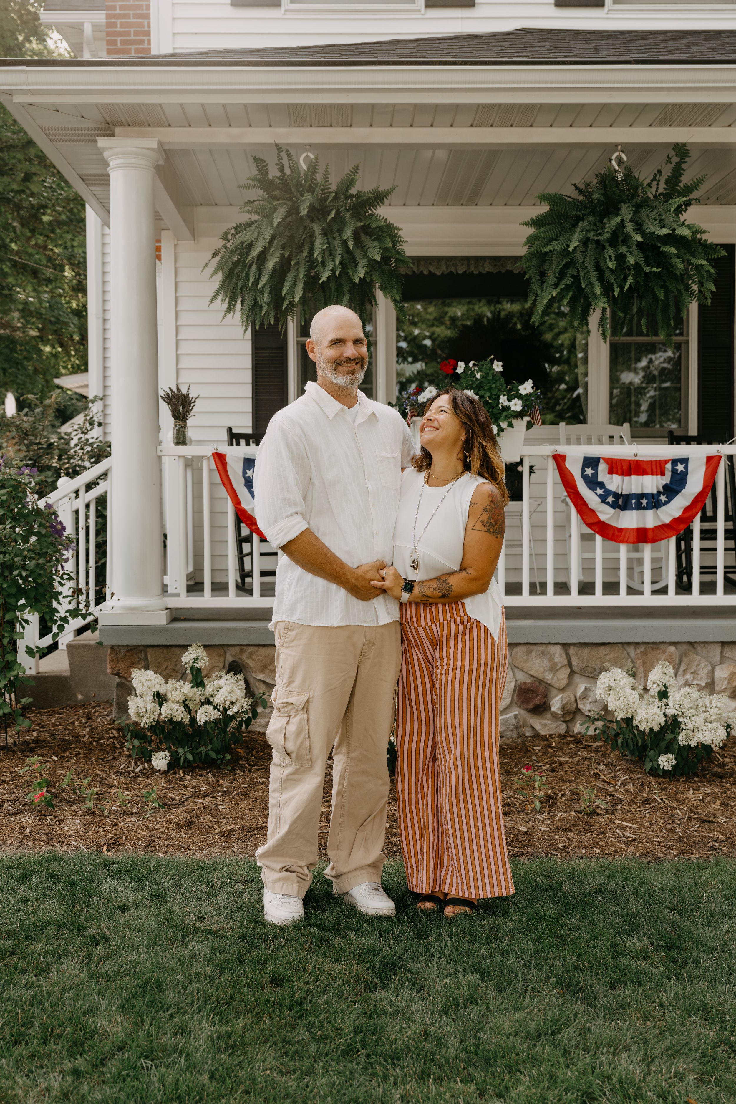 Couple poses in front of white house porch with American bunting and hanging ferns for a patriotic summer photo.
