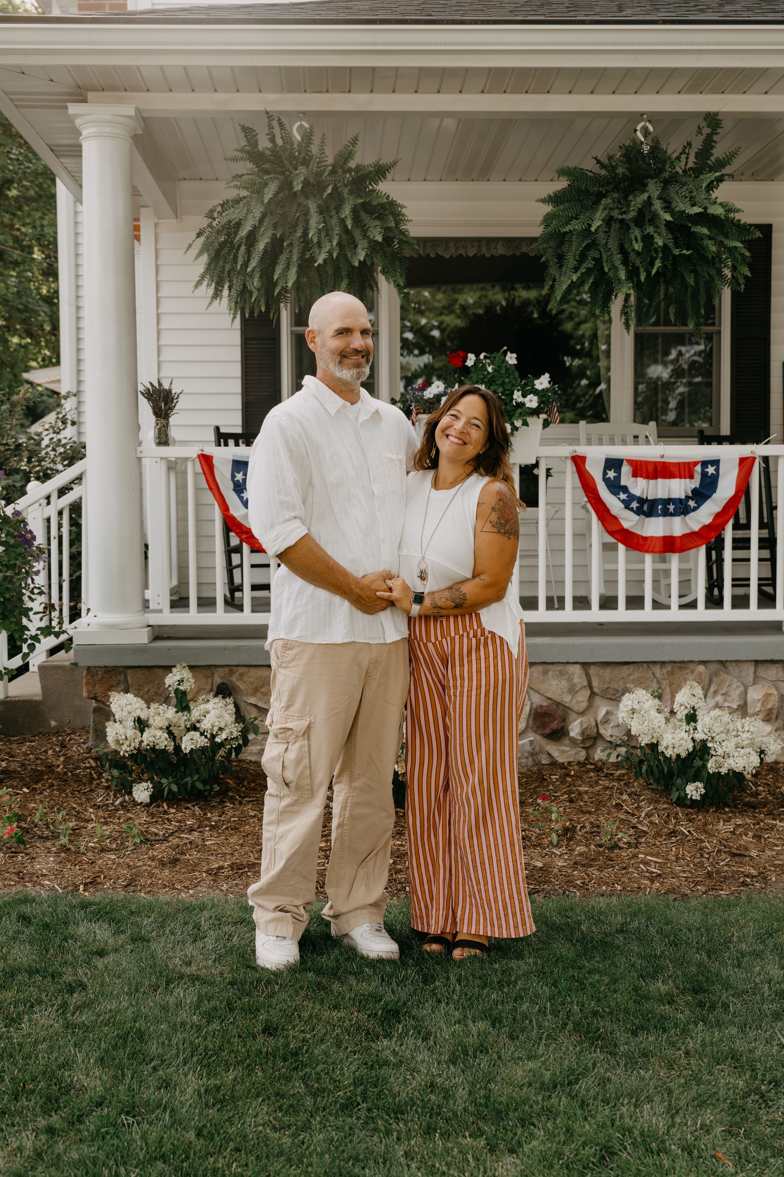 A couple stands on their front porch decorated with patriotic bunting and ferns on a summer day.